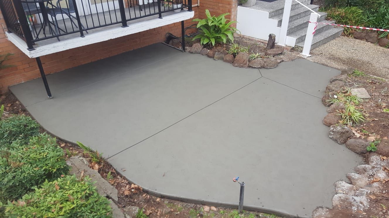 Freshly poured concrete patio beside a house, bordered by garden beds and a deck railing