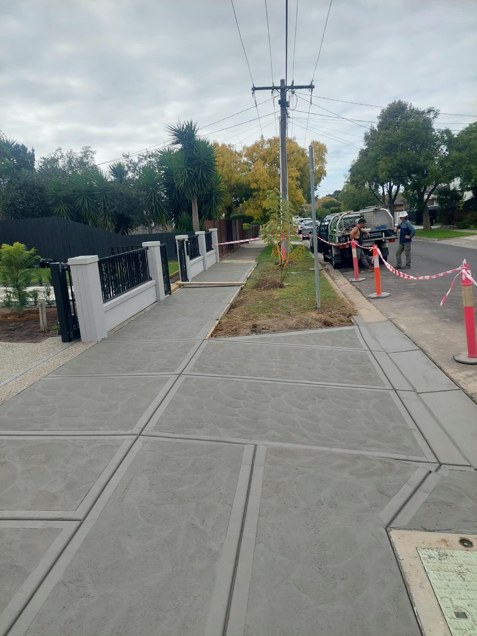 Sidewalk beside a fenced street with a utility pole, trees, and orange traffic cones blocking part of the path.