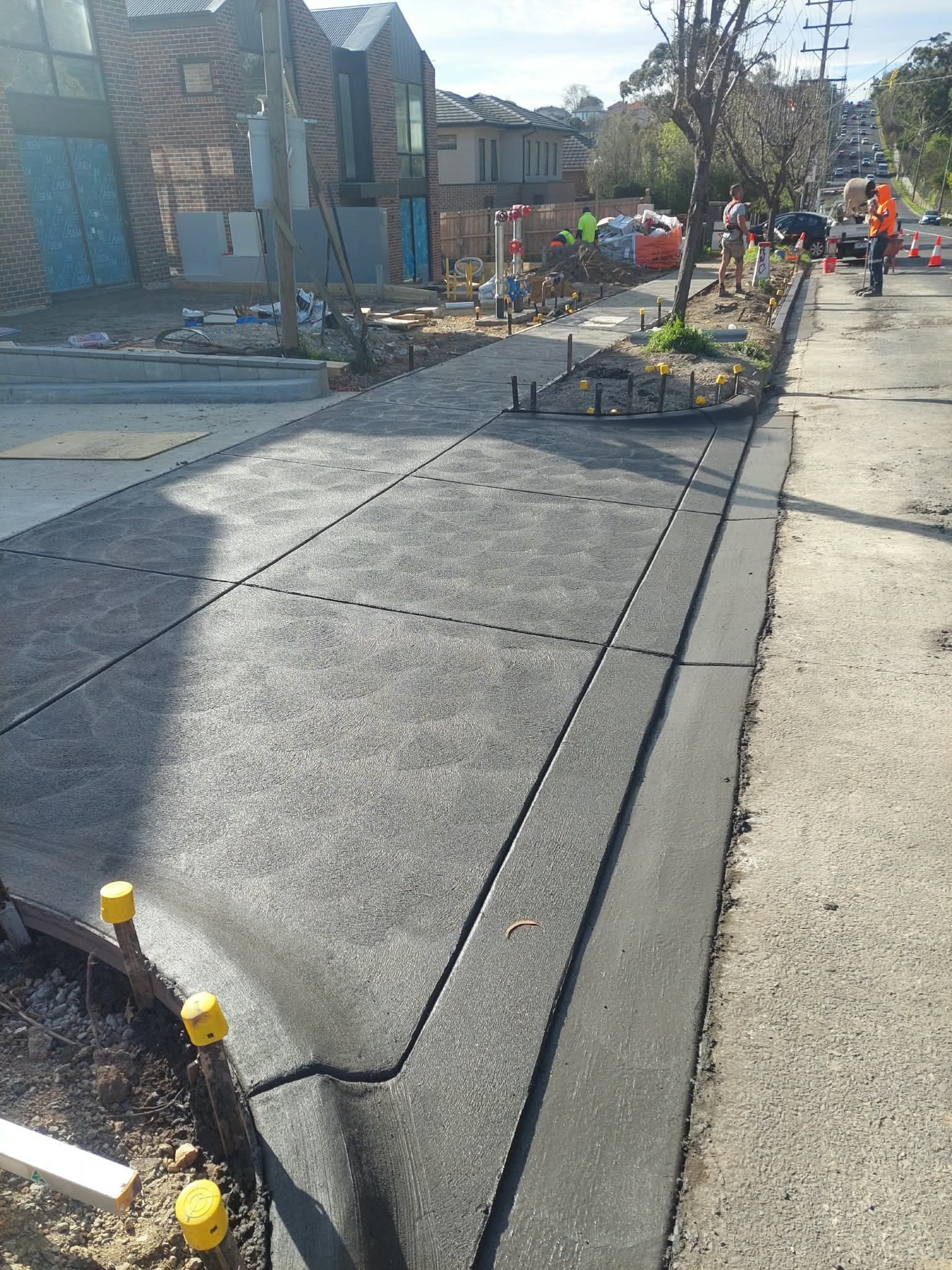 Workers repairing a cracked sidewalk beside a street, with barriers and construction cones
