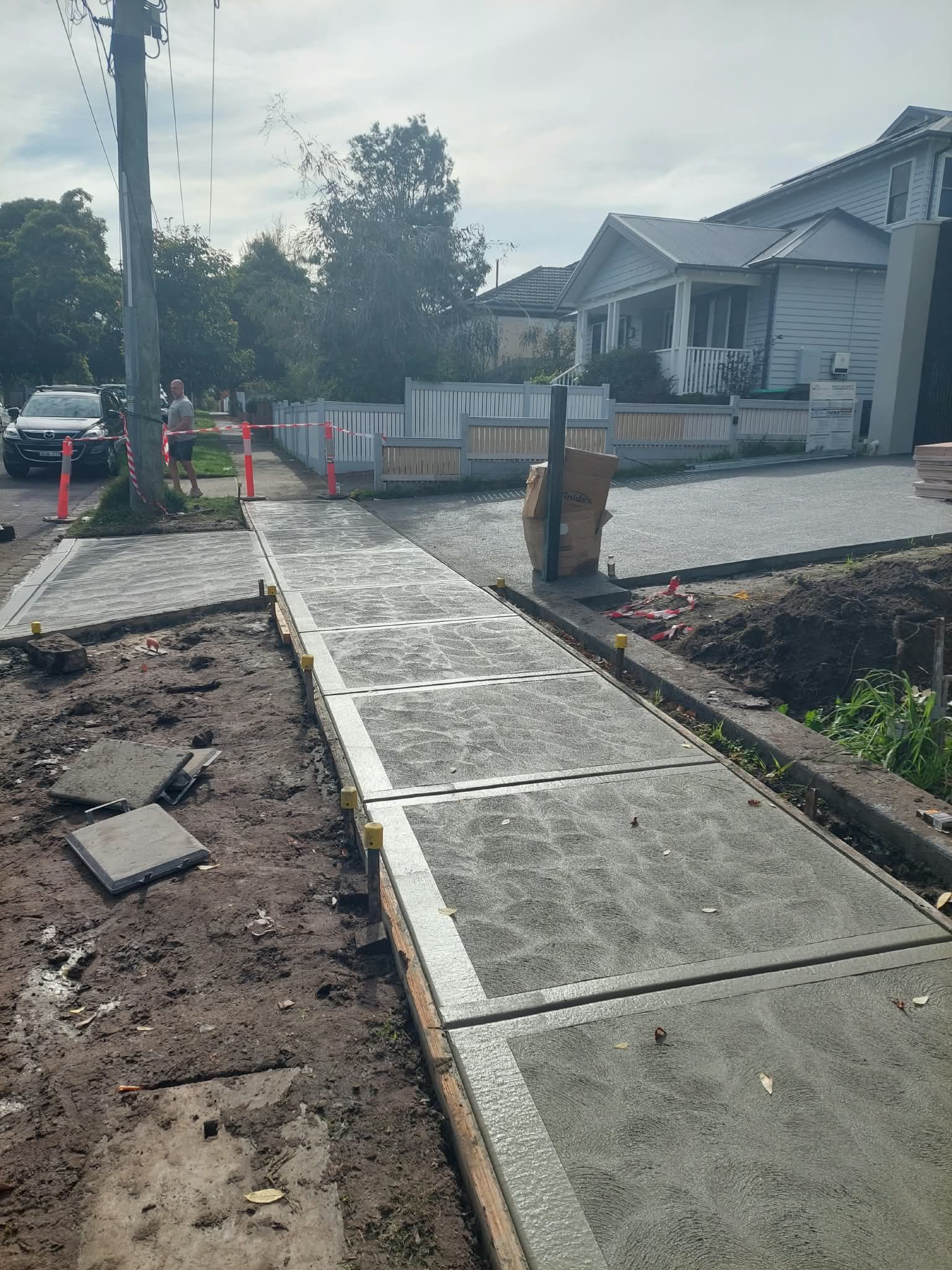 Sidewalk under construction beside homes, with fresh concrete panels, dirt edges, and orange safety cones.