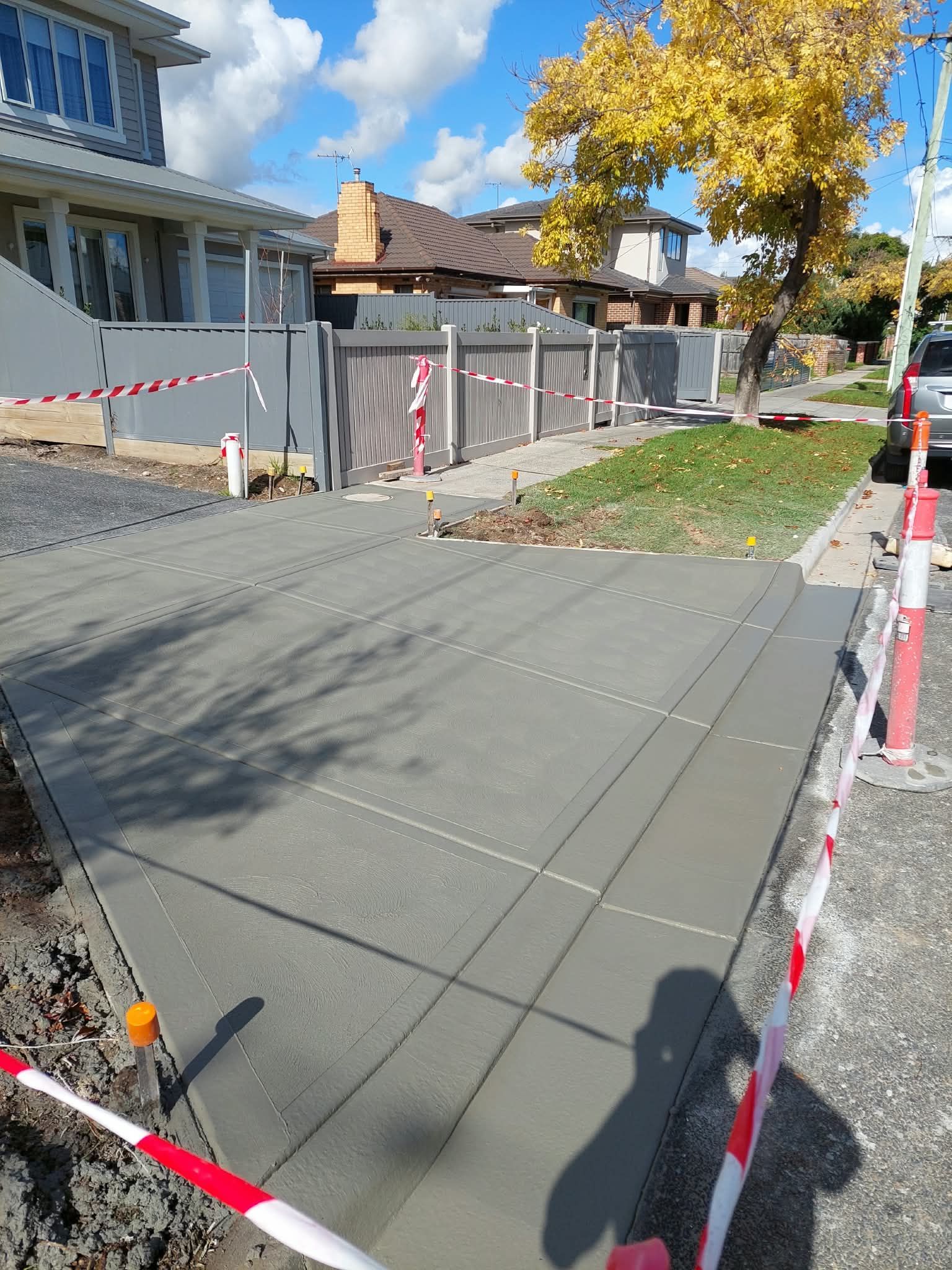 Cement sidewalk under construction beside houses, bordered by red-and-white safety tape and orange cones.