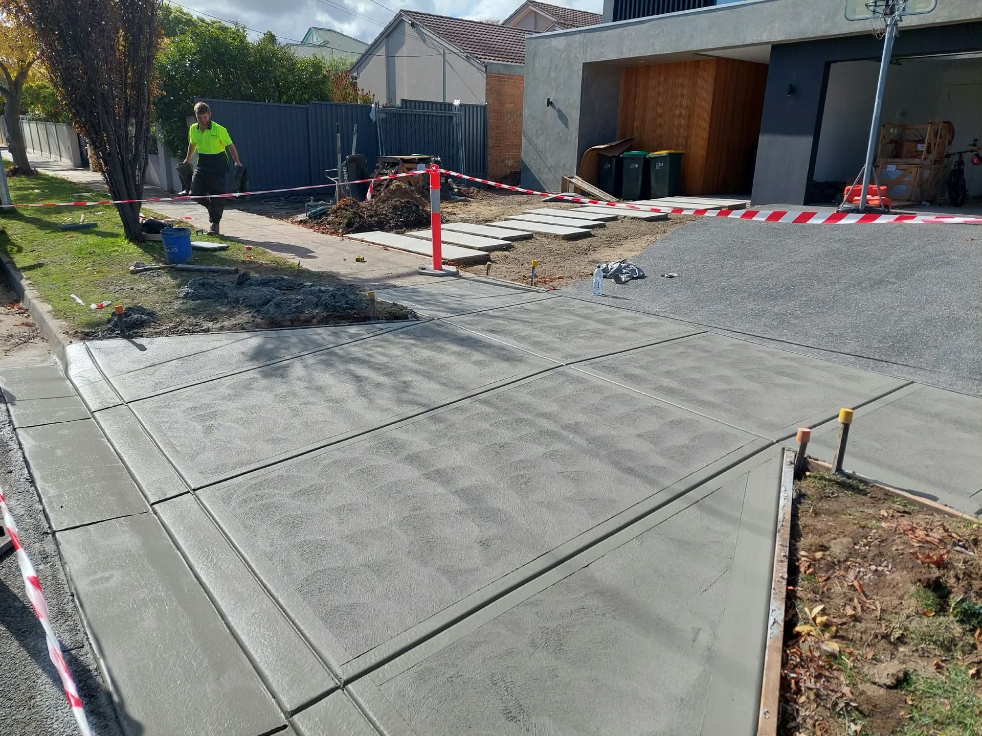 Concrete driveway and sidewalk under construction beside a house, with red-and-white caution tape and workers in background