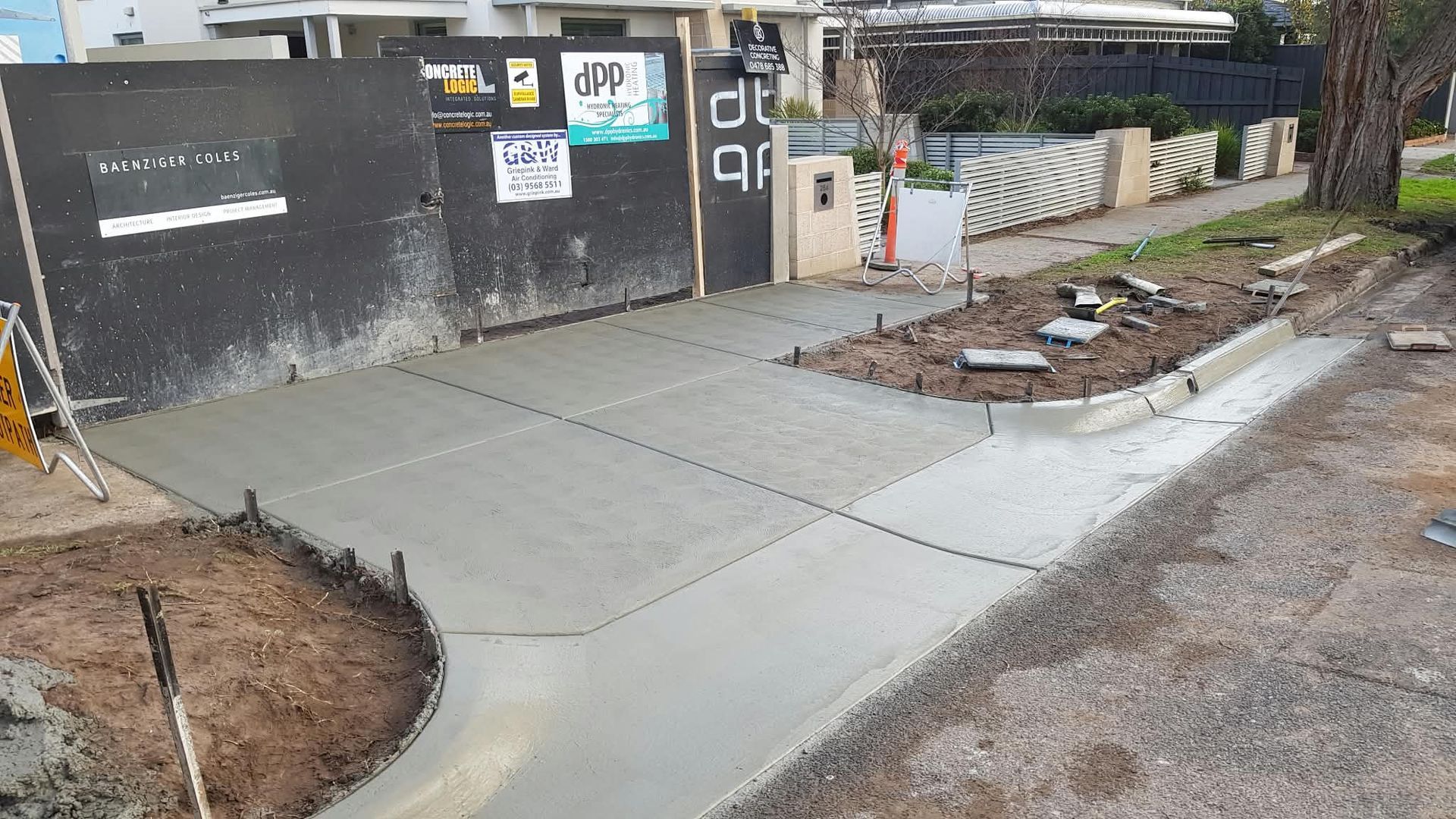 Concrete sidewalk path beside a black wall and gate, with muddy ground and a small landscaped strip.