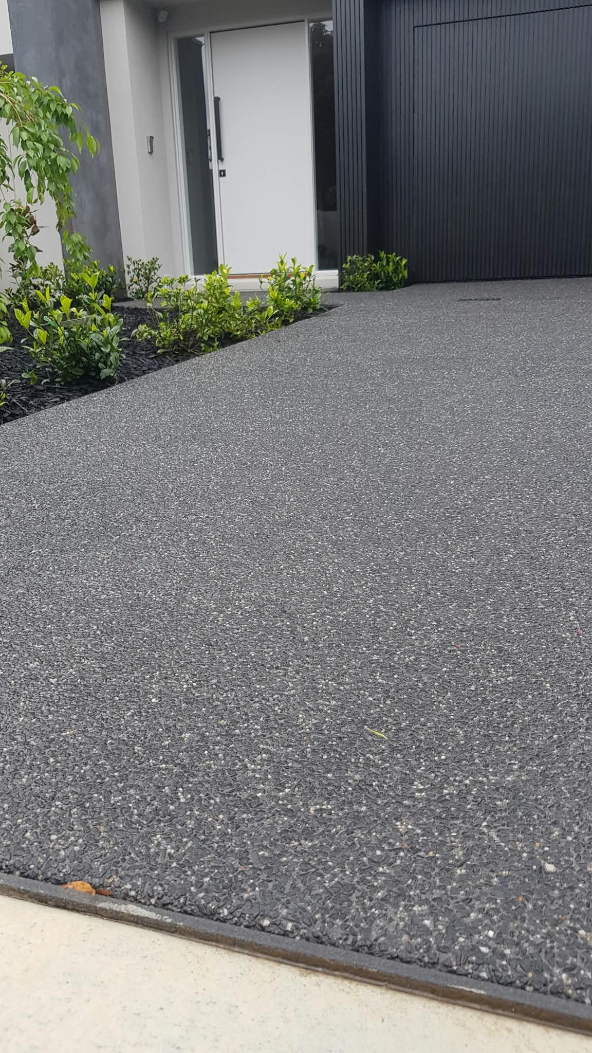 Gravel driveway beside a white wall, black gate, and small green shrubs
