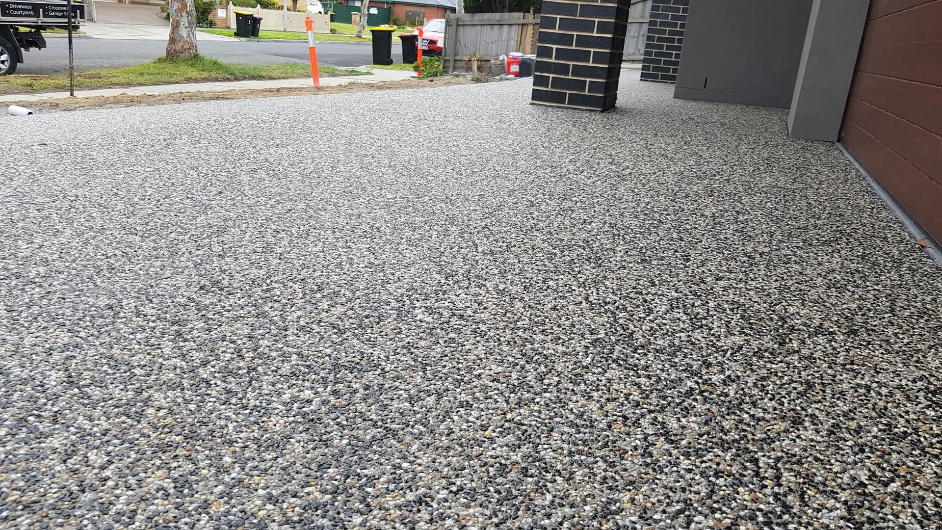 Freshly poured speckled concrete driveway beside a house, with street and traffic cones in the background