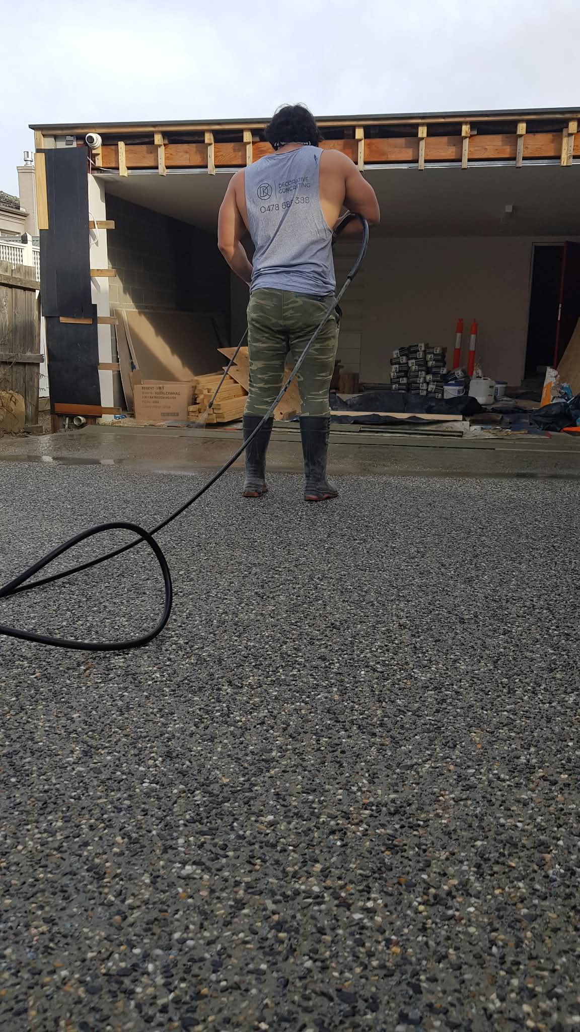 Person pressure-washing a gravel driveway in front of a partially destroyed garage, with debris and exposed roof beams.