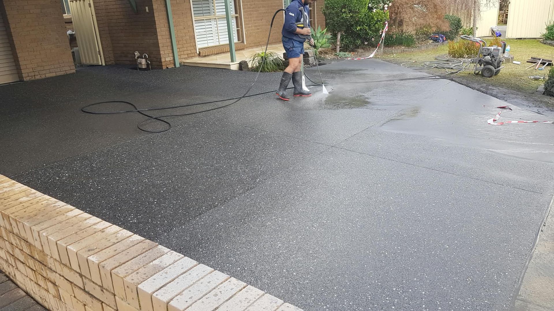 Person pressure-washing a wet black driveway beside a brick house.