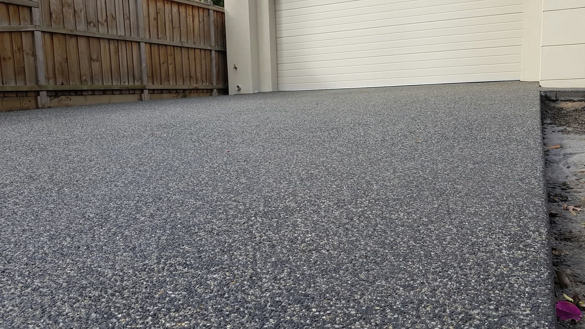 Gravel driveway leading to a white garage door beside a wooden fence.