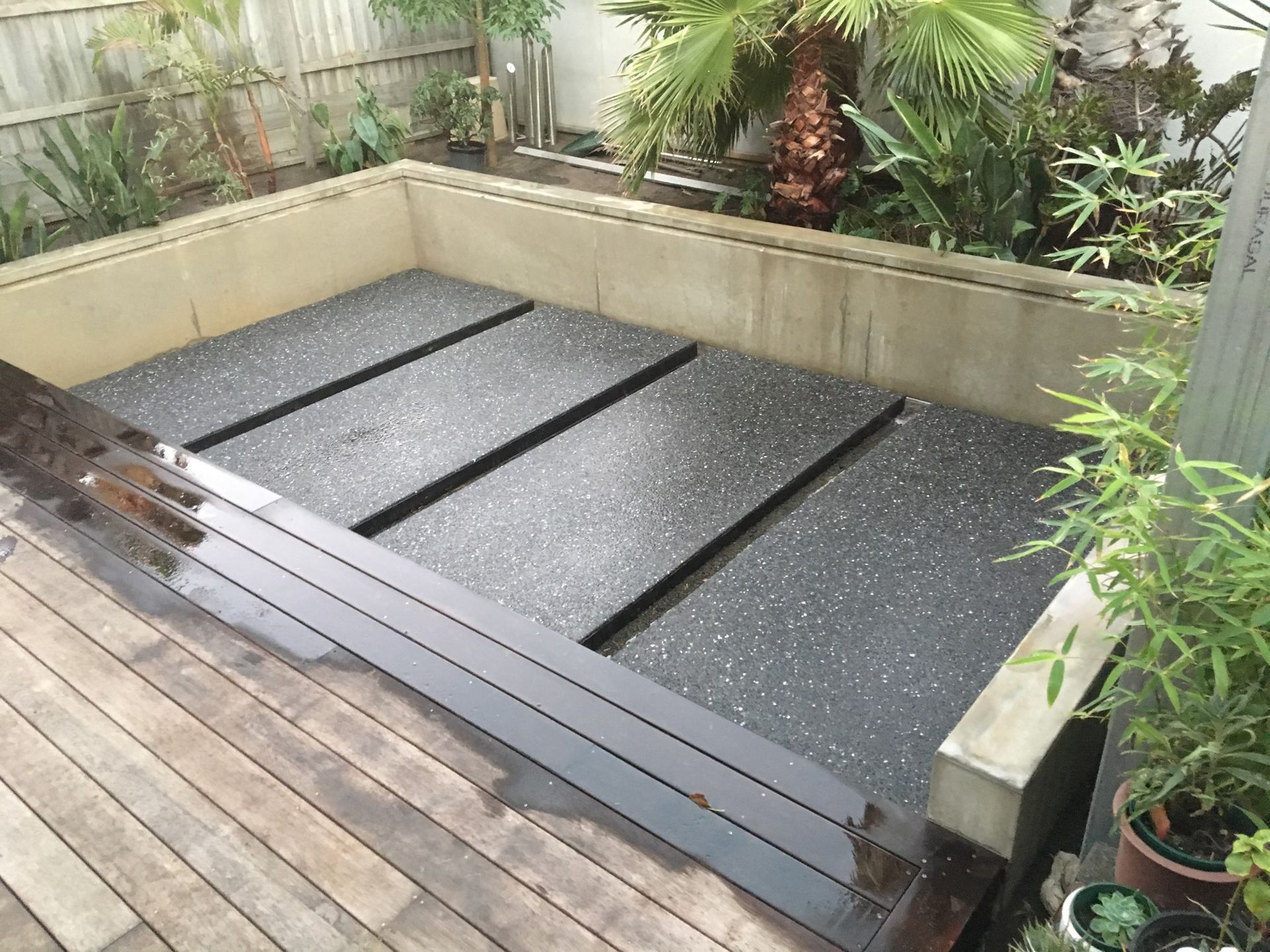 Raised backyard planter with black gravel and dark wooden planks beside a timber deck, bordered by plants