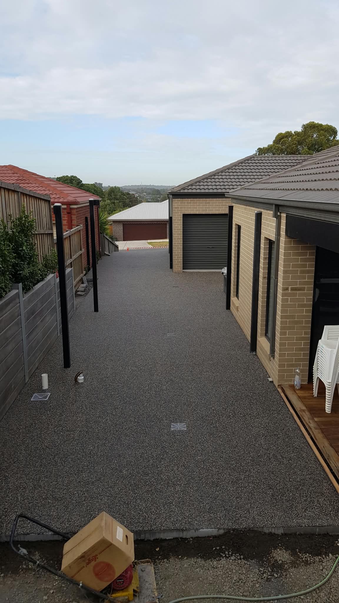 Gravel driveway between two houses, with a cloudy sky and a cardboard box in the foreground.