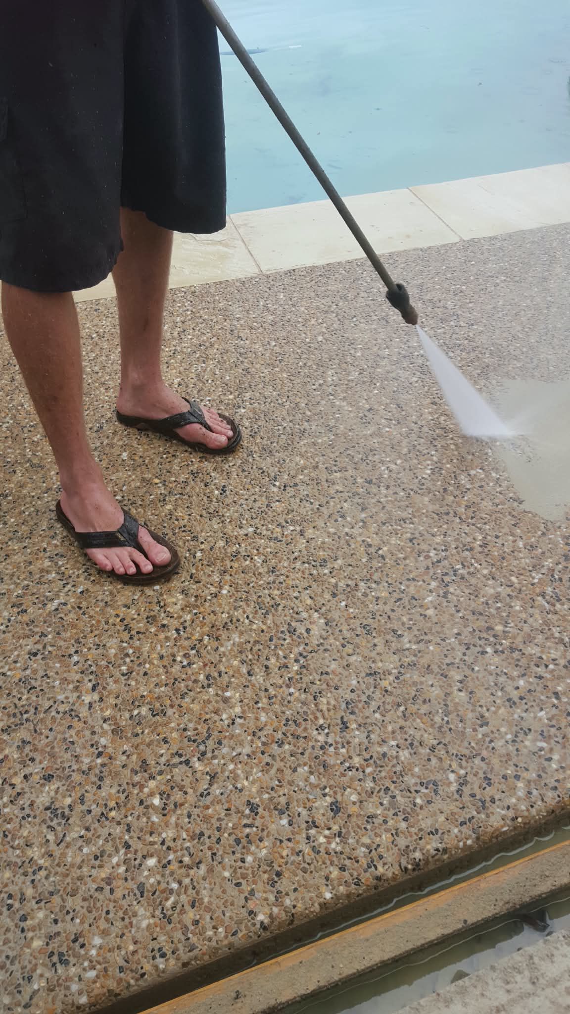 Person using a pressure washer on a pebble surface beside a pool