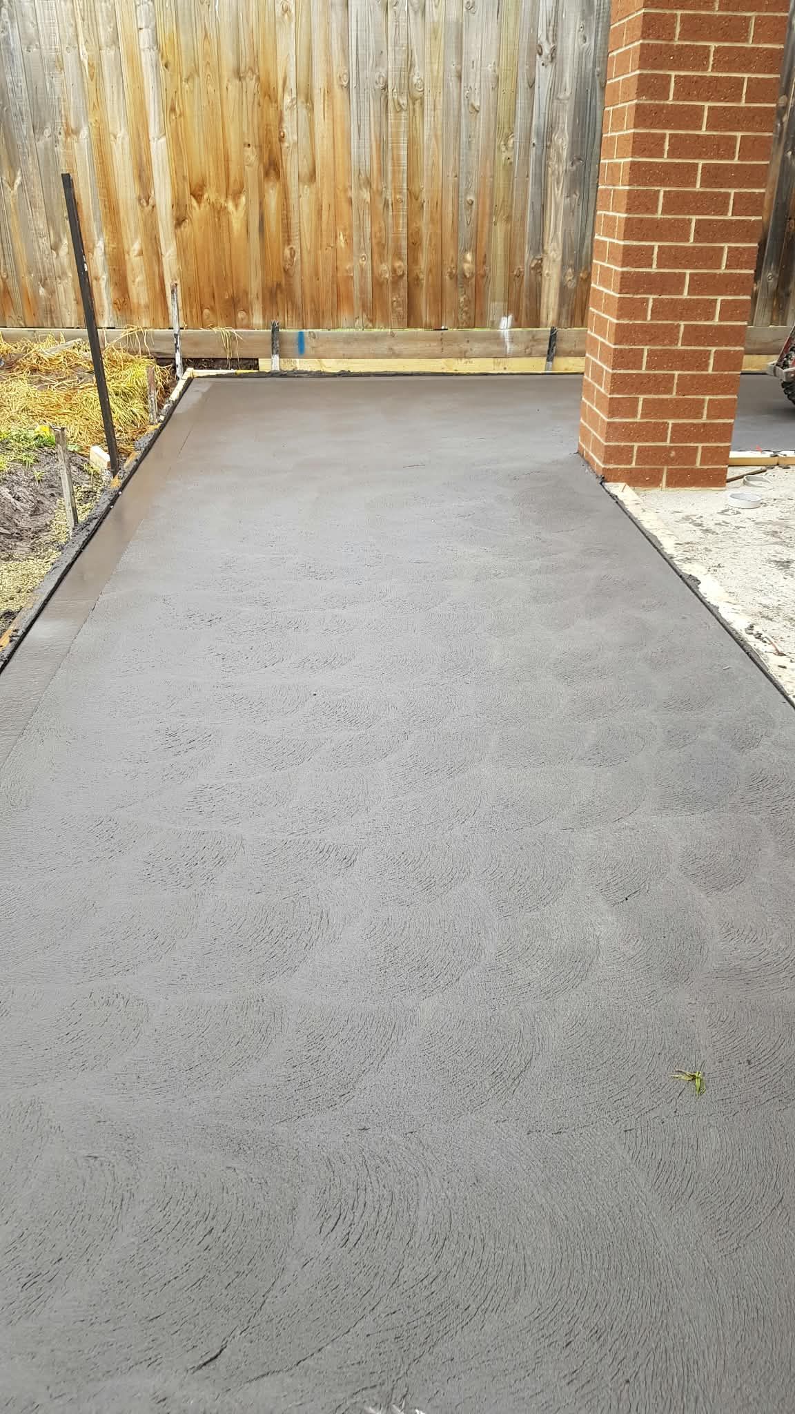 Freshly paved dark concrete walkway beside a brick pillar and wooden fence.