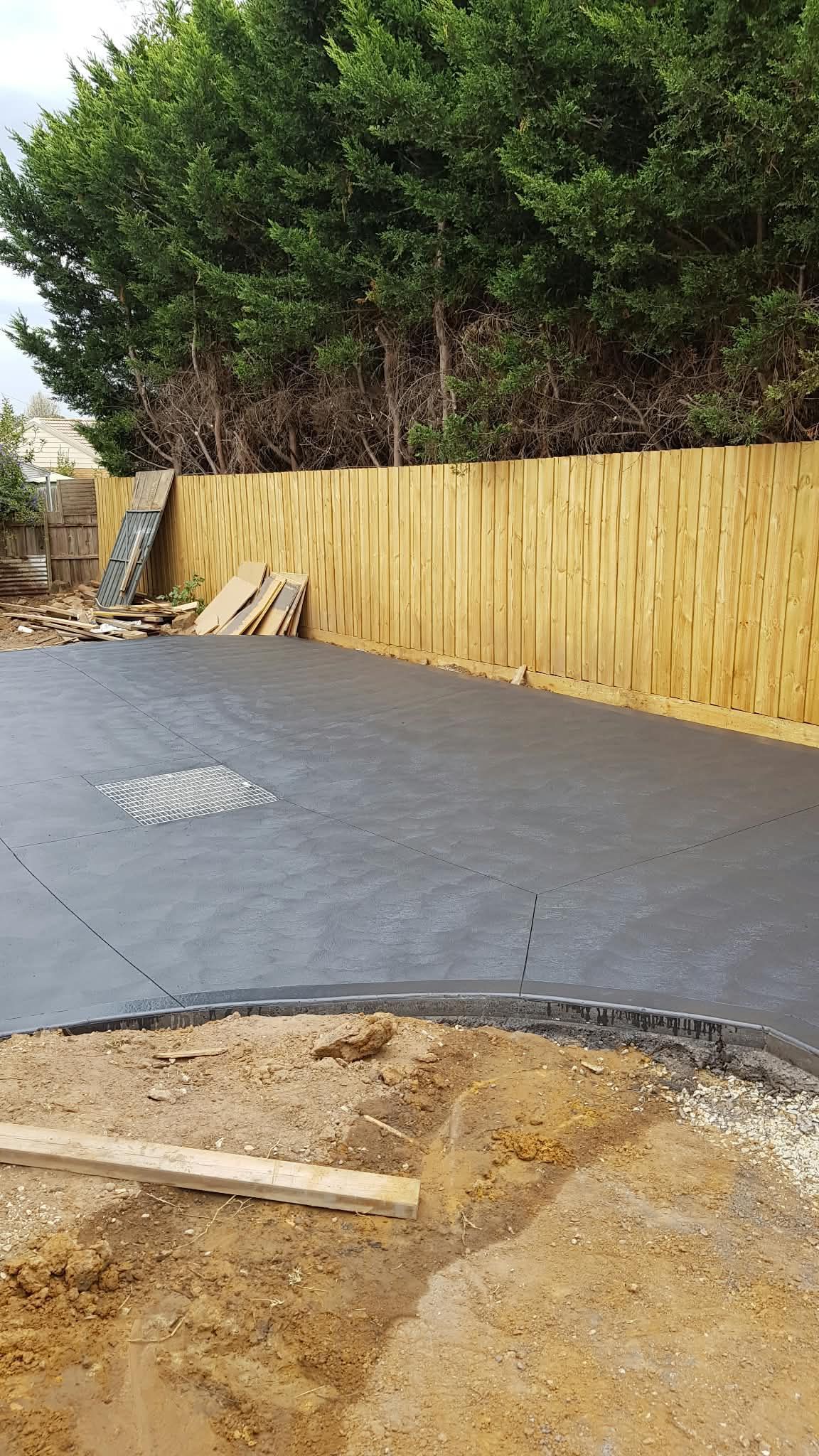 New concrete driveway beside a wooden fence, with trees in the background and unfinished dirt foreground