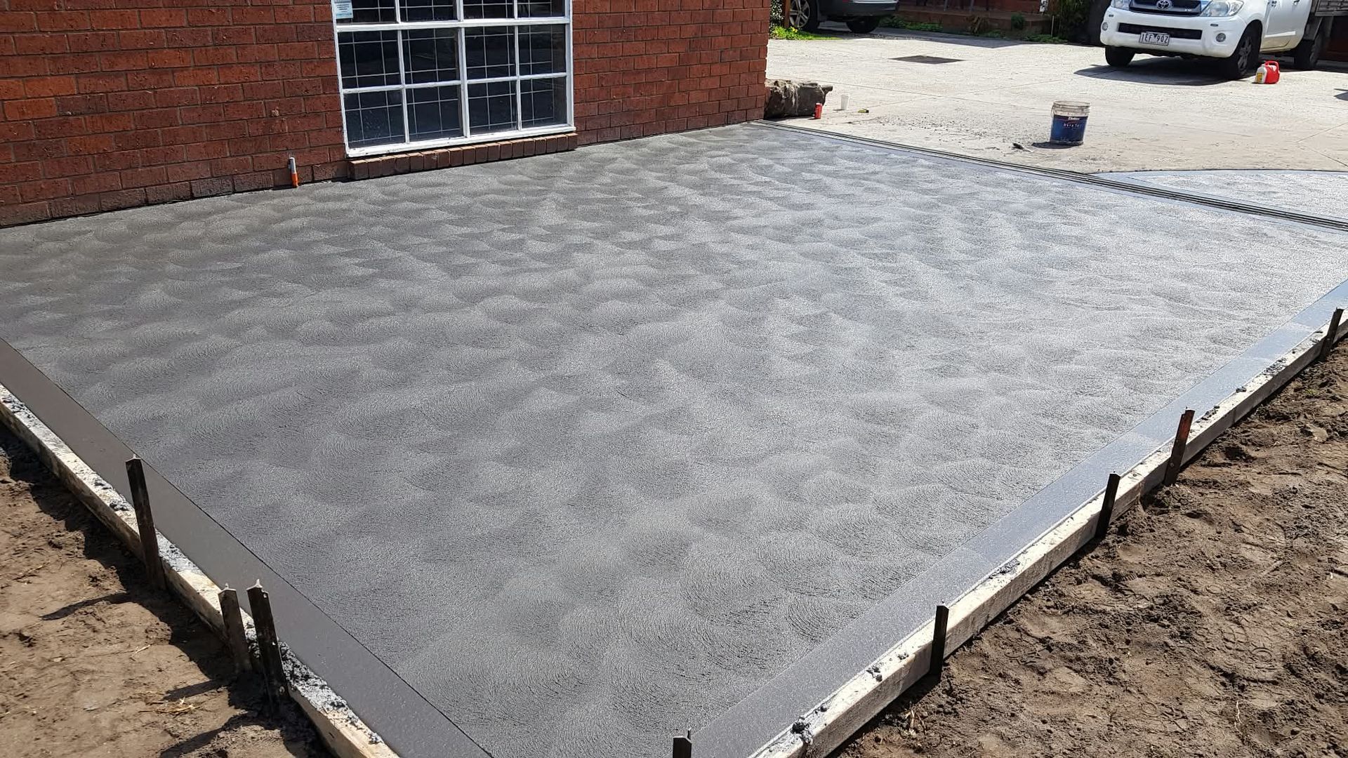 New concrete patio slab beside a brick house, bordered by wooden forms and dirt.