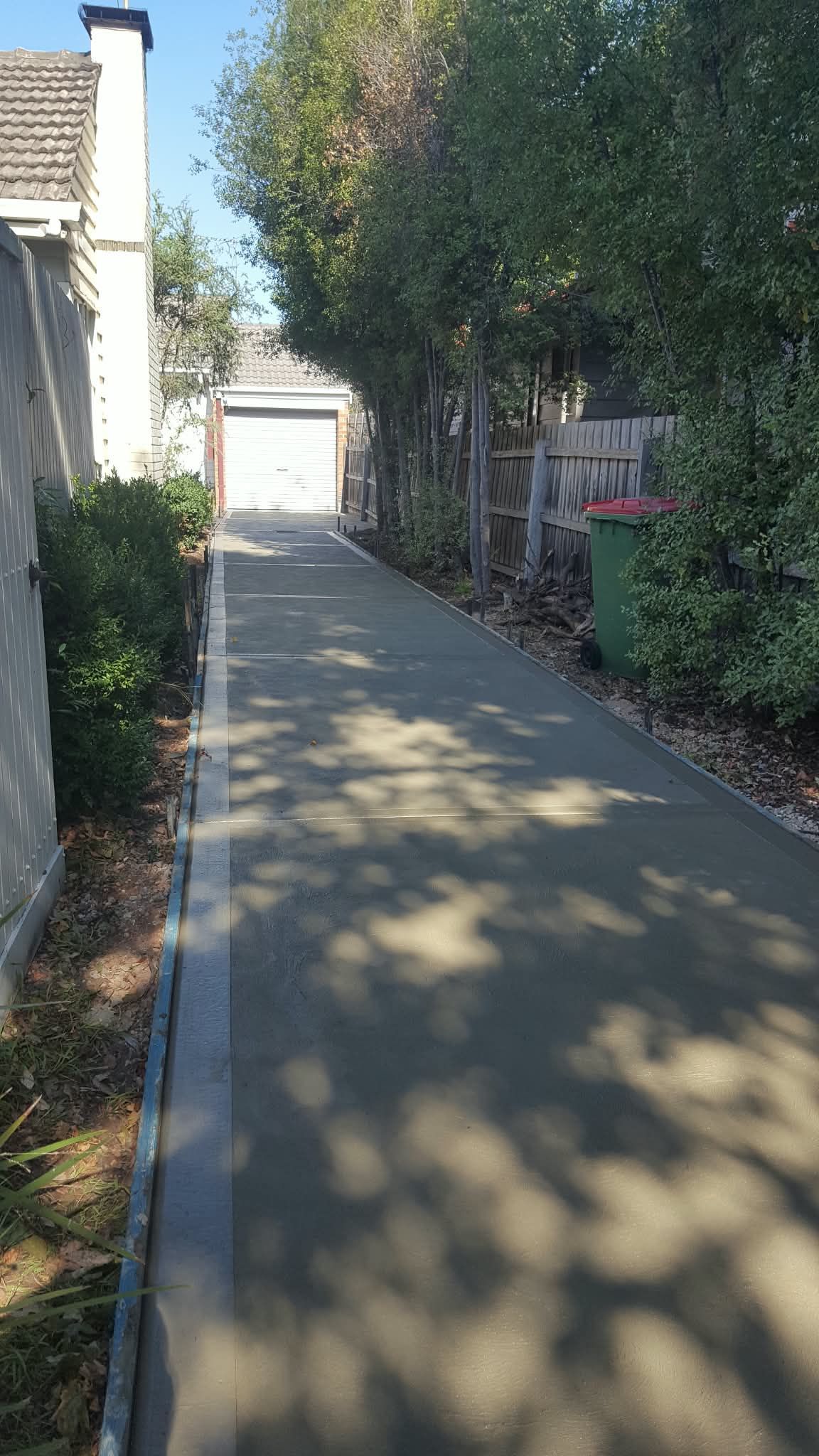 Narrow paved driveway beside a house, lined with trees and fences, leading to a closed garage.