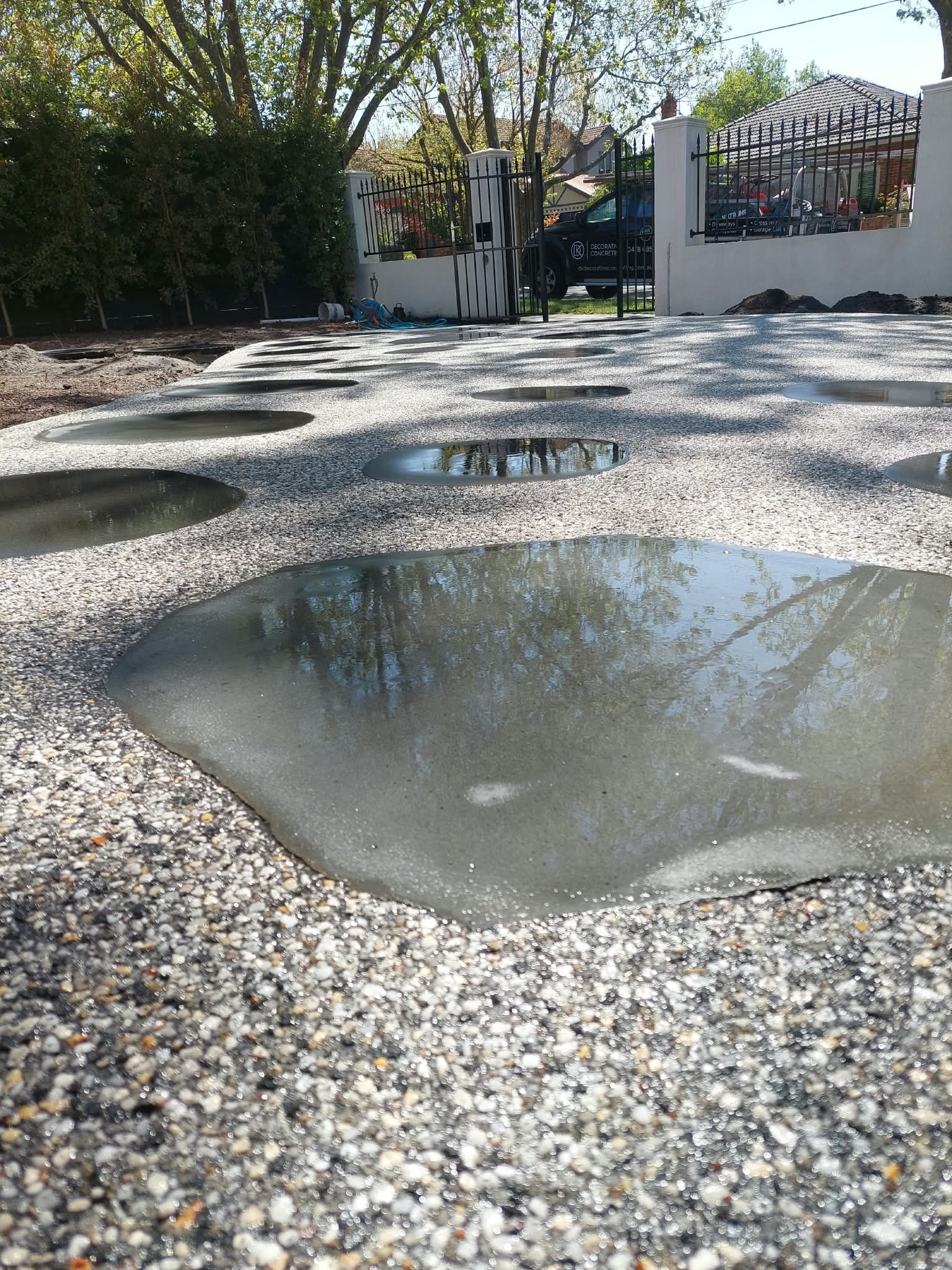 Puddles of water on a gravel path by a garden gate and trees on a sunny day
