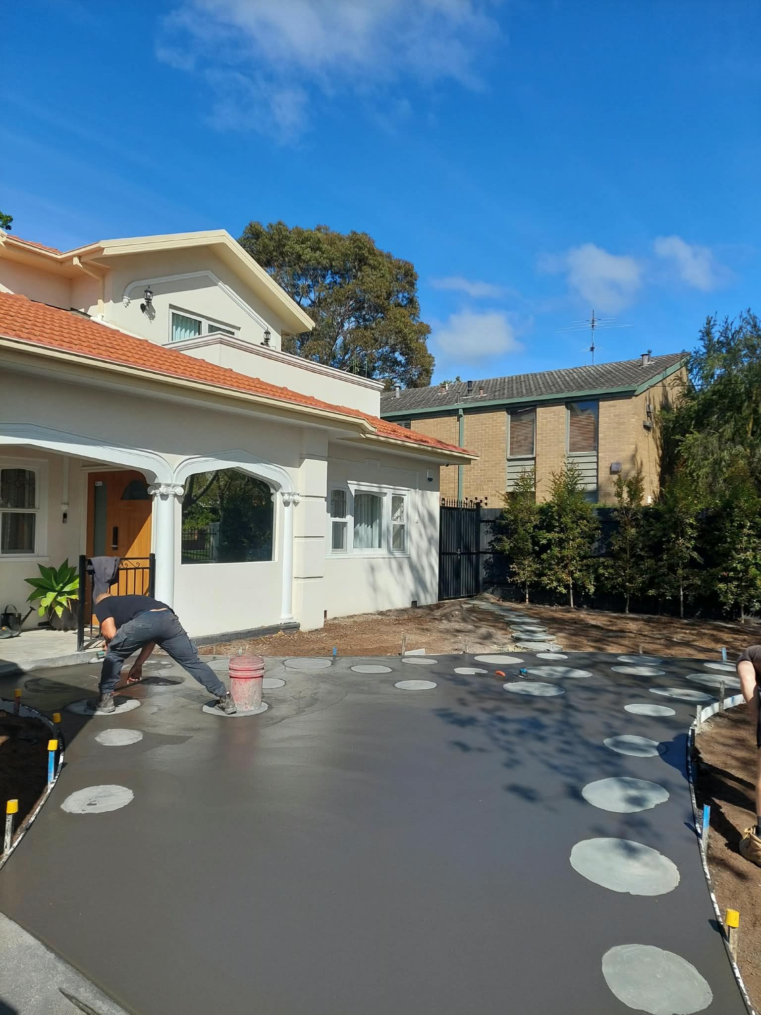 Worker smoothing fresh gray concrete patio beside a white house under a blue sky