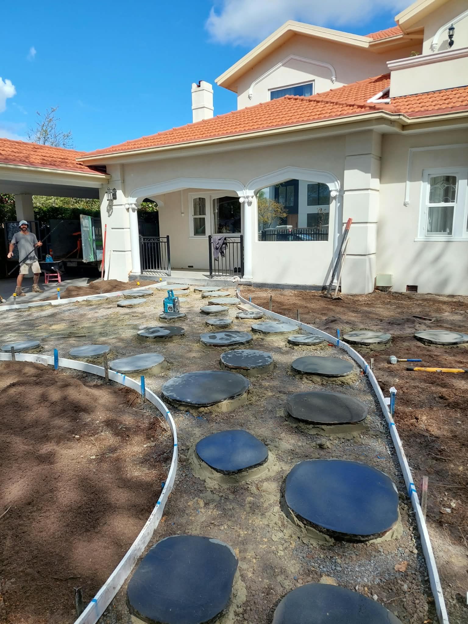 Backyard patio under construction with stepping stones, hoses, and a white house with red tile roof.