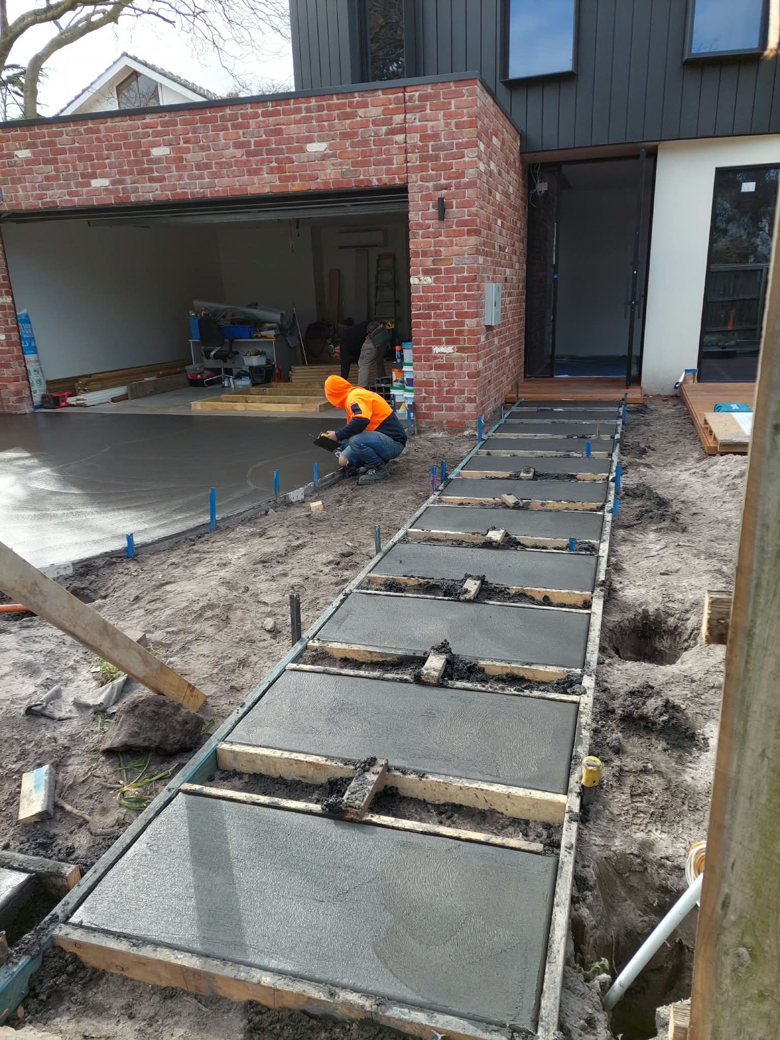 Worker pours concrete in a driveway beside a brick garage under construction.