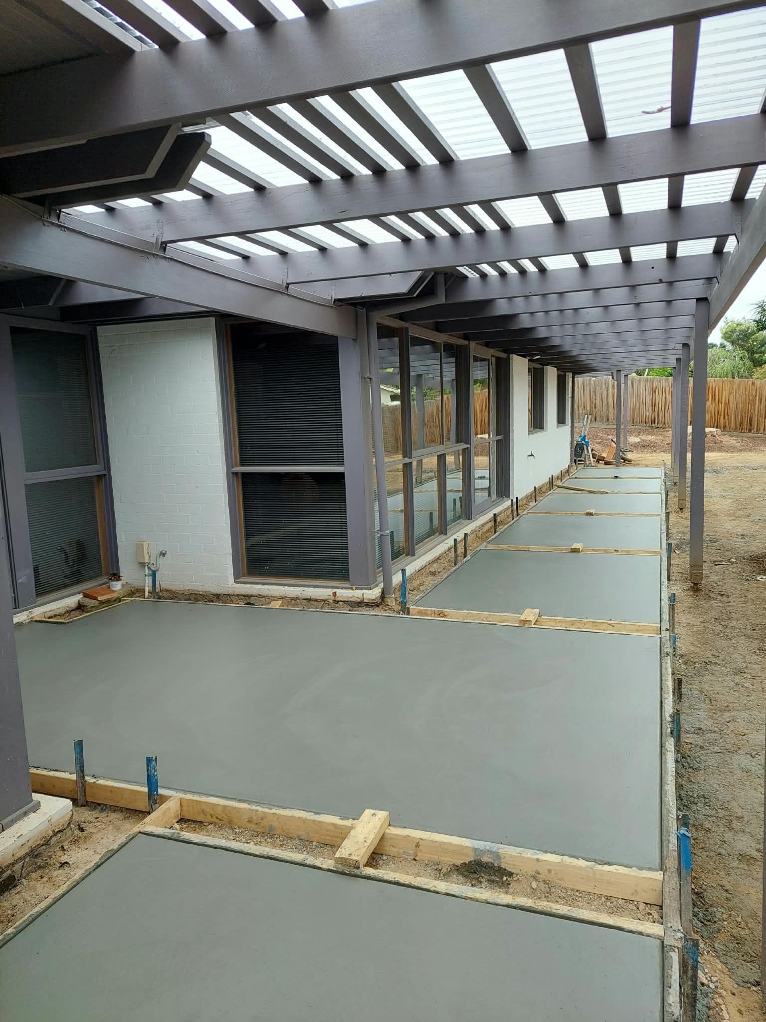 Covered outdoor walkway with fresh concrete slabs, wooden forms, and a slatted overhead roof