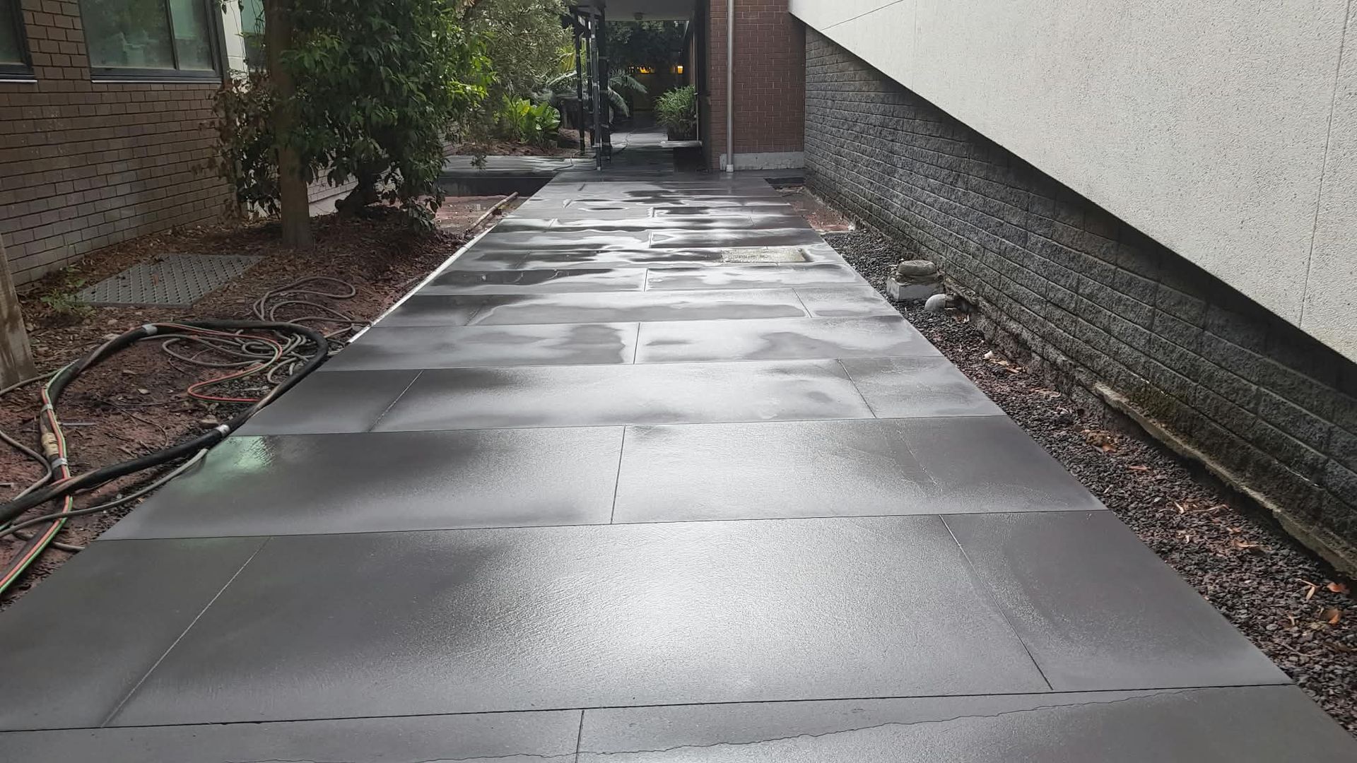 Wet concrete walkway leading between a brick wall and a white building toward a doorway