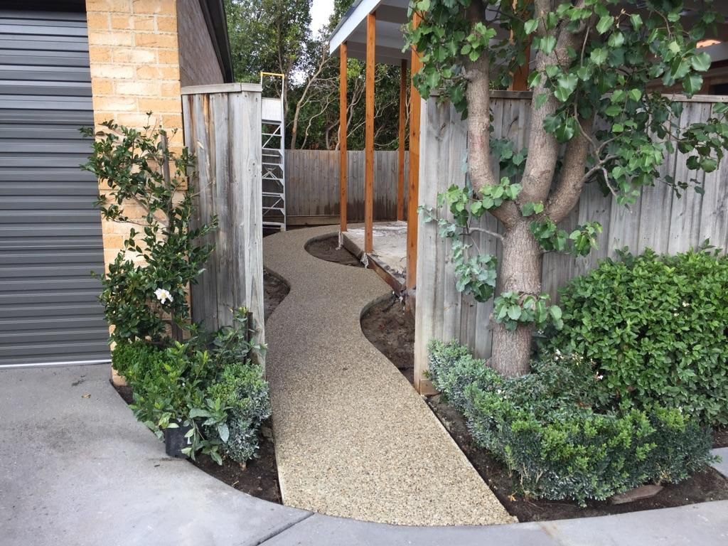 Narrow gravel garden path between a garage and vine-covered fence, with wooden posts and greenery