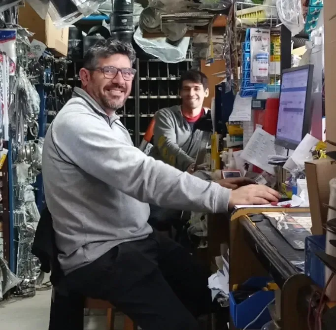 Dos personas sonriendo a un mostrador de tienda desordenado, con estantes y artículos colgados apilados a su alrededor.