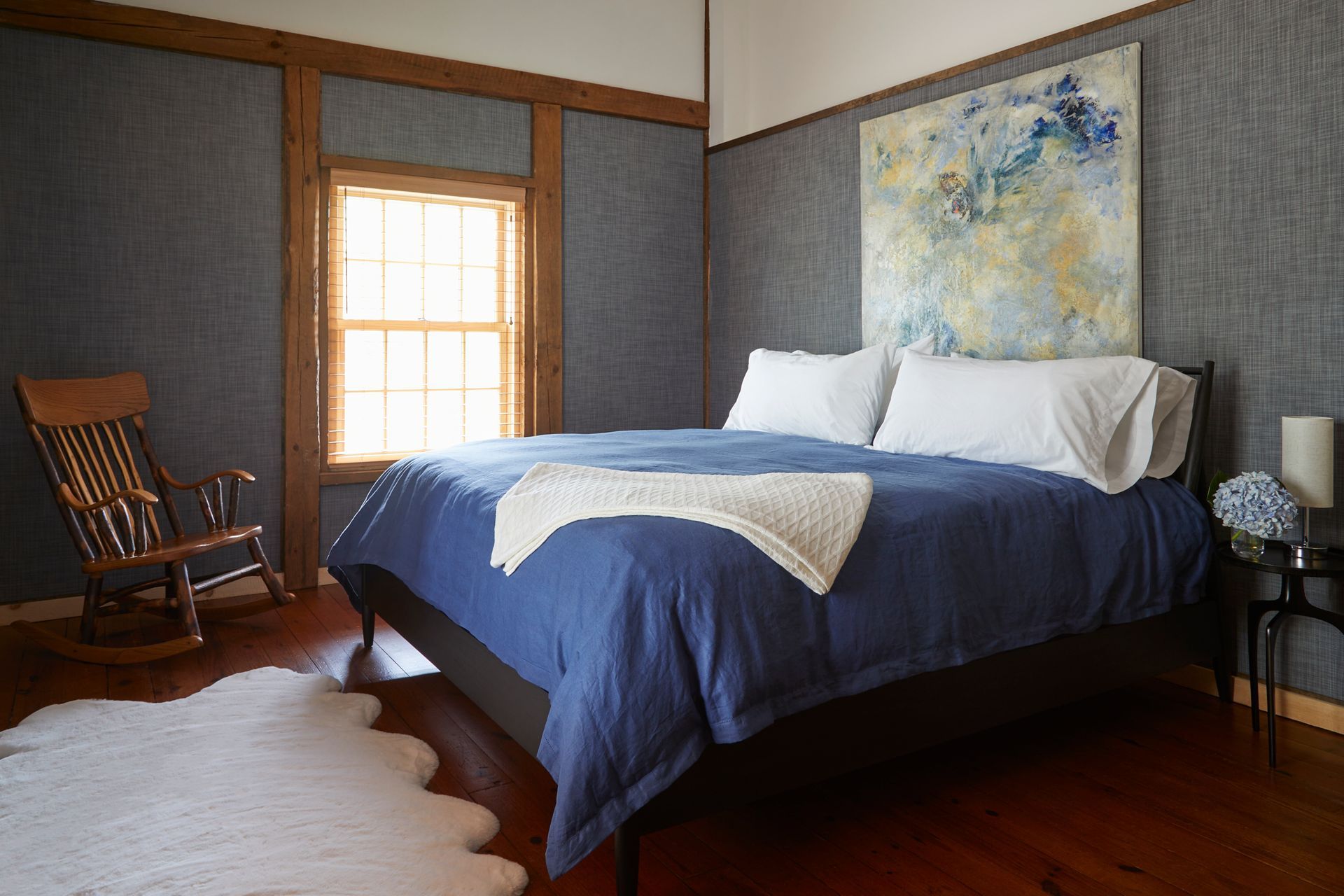 Bedroom with a blue bedspread, white pillows, wooden chair, and sunlight through a window.