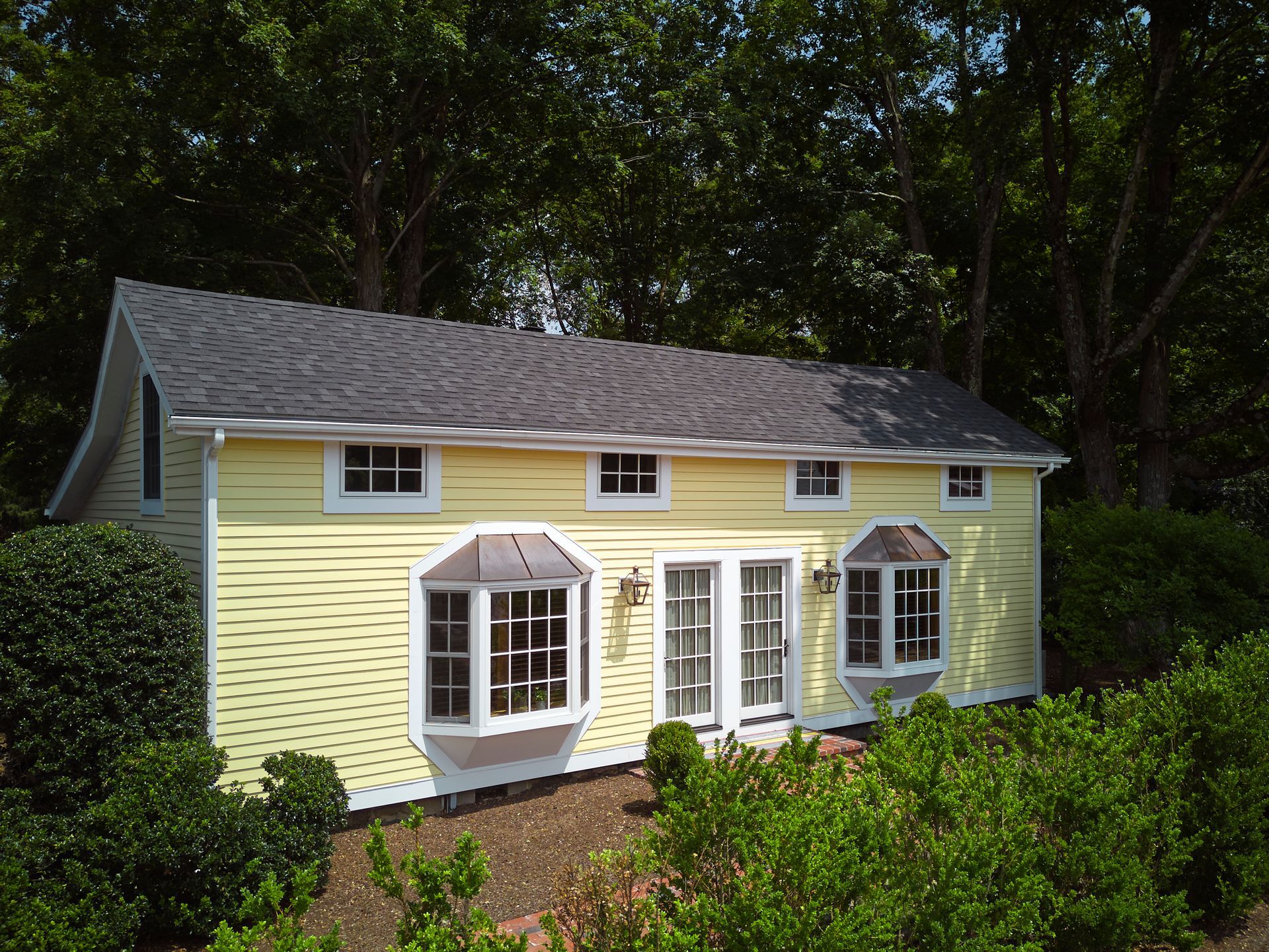Yellow cottage with gray roof, white trim, and bay windows surrounded by green shrubs and trees