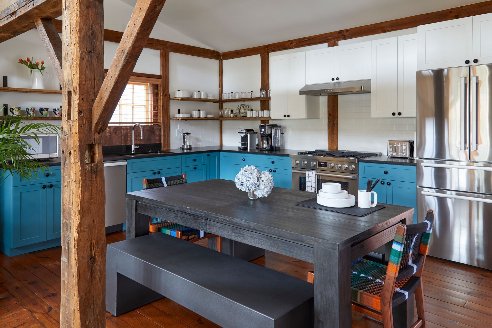 Rustic kitchen with blue cabinets, wooden beams, and a dark dining table in the foreground