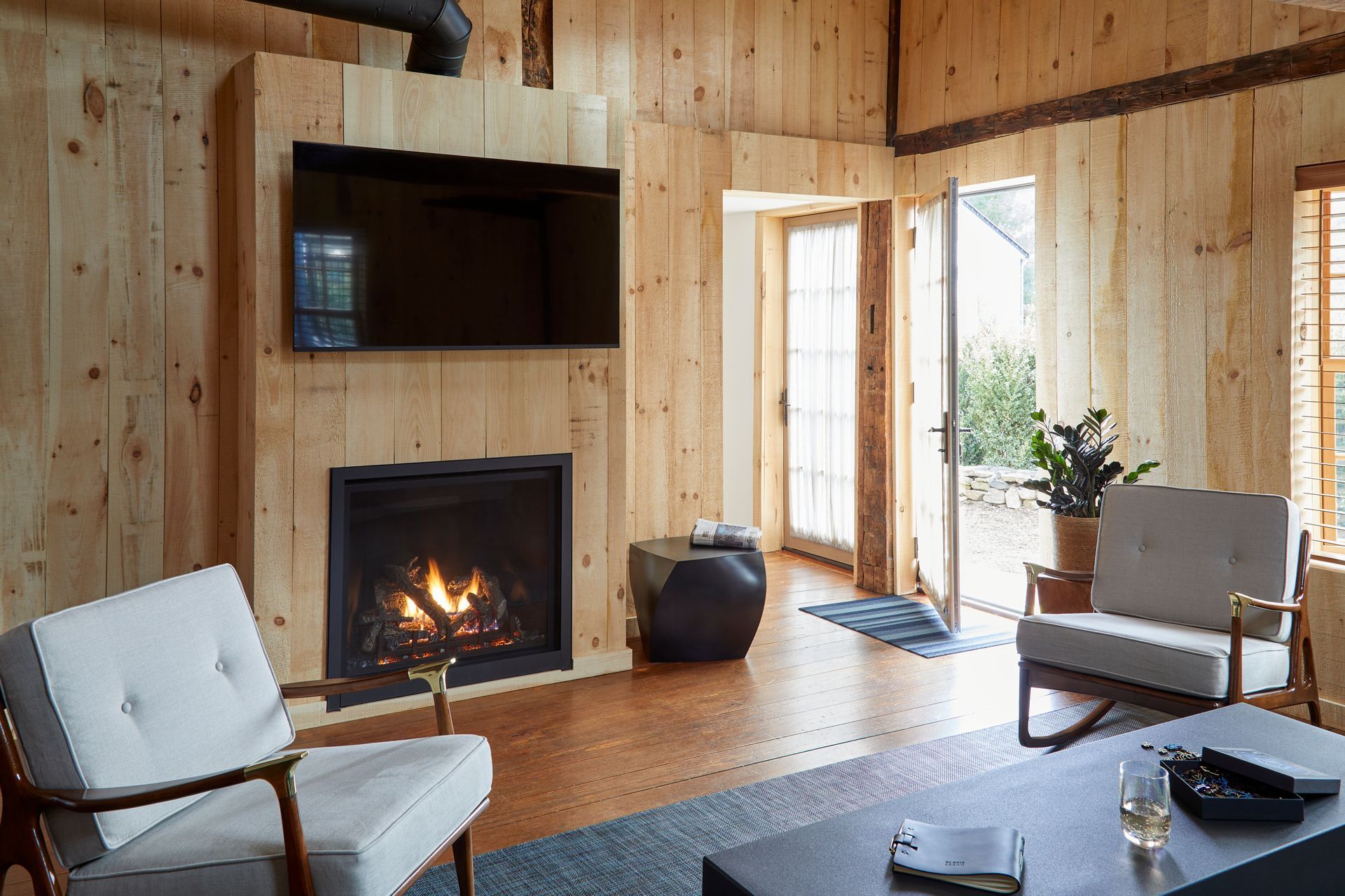 Wood-paneled living room with fireplace, wall-mounted TV, and chairs by a bright window.