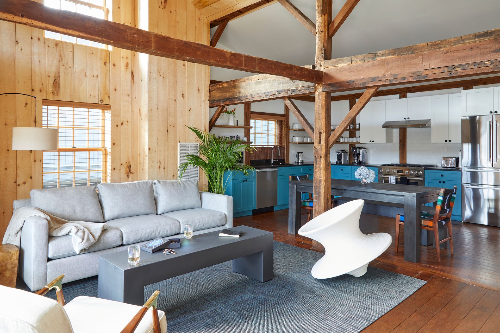 Open loft living room with gray sofa, blue kitchen, wood beams, and dining area