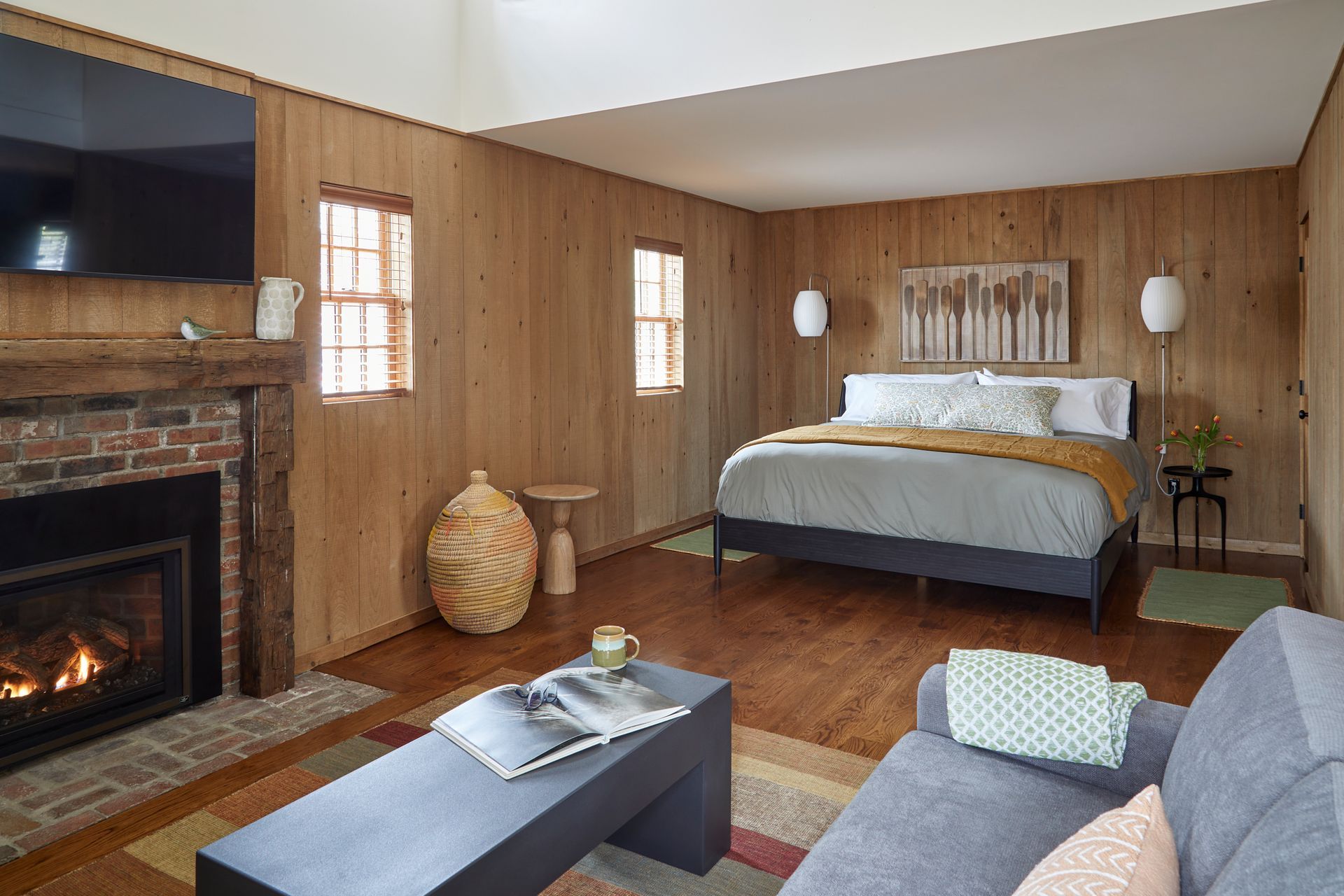 Cozy bedroom with wood-paneled walls, fireplace, queen bed, and gray sofa seating area