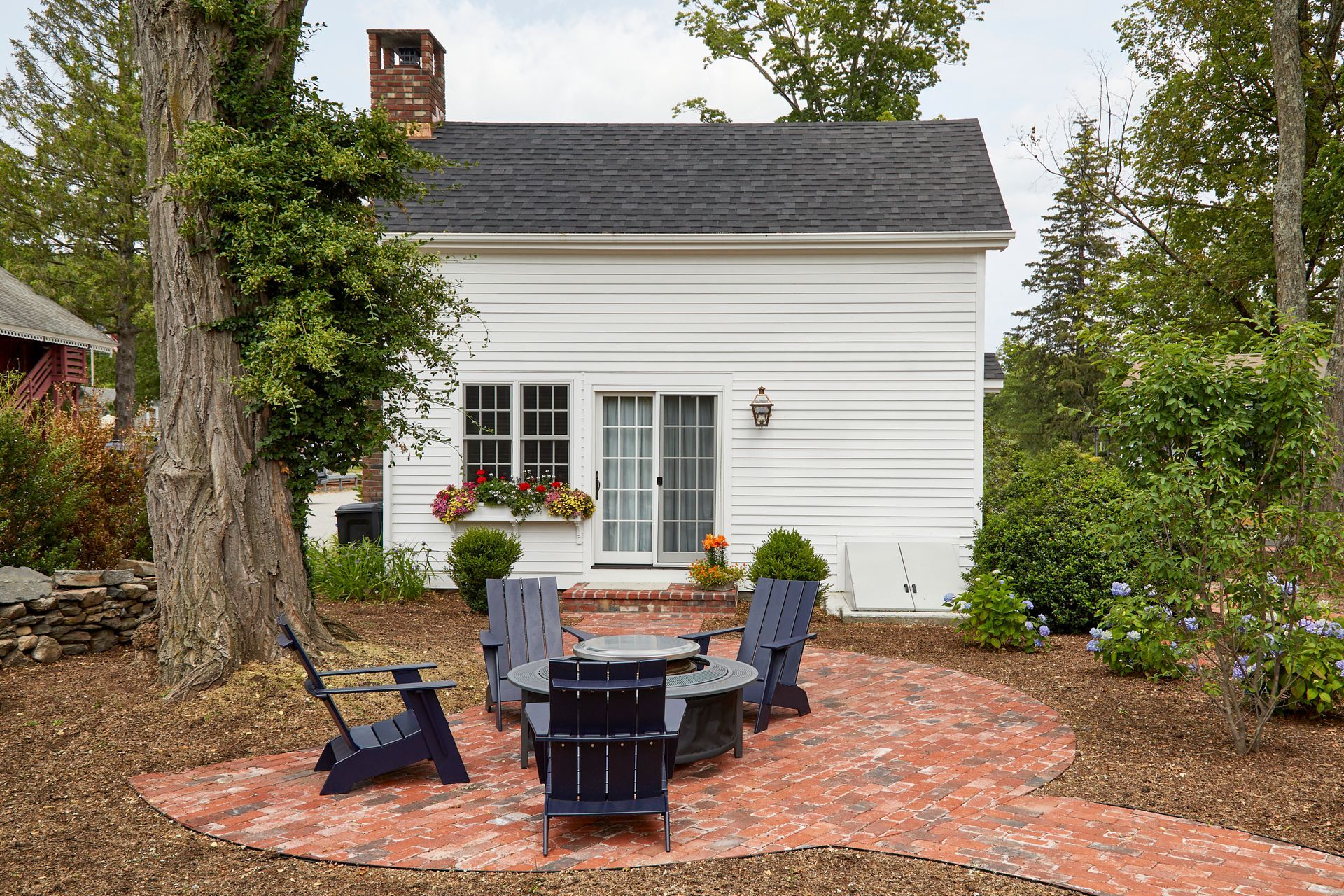 White cottage with a brick patio, black chairs, and a round table in a wooded yard
