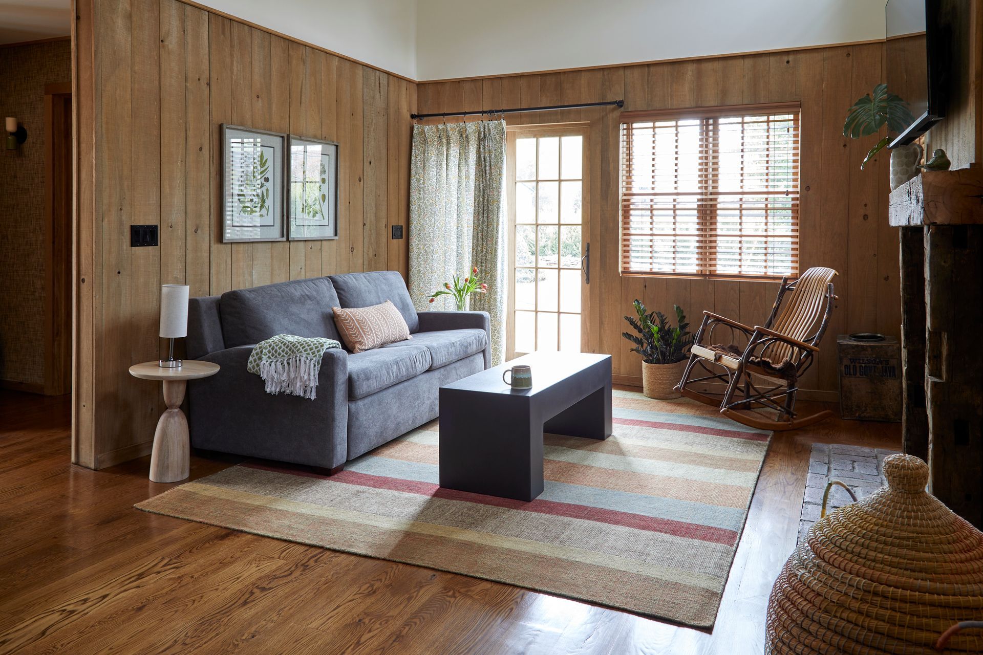 Wood-paneled living room with gray sofa, coffee table, rocking chair, and large windows with blinds.