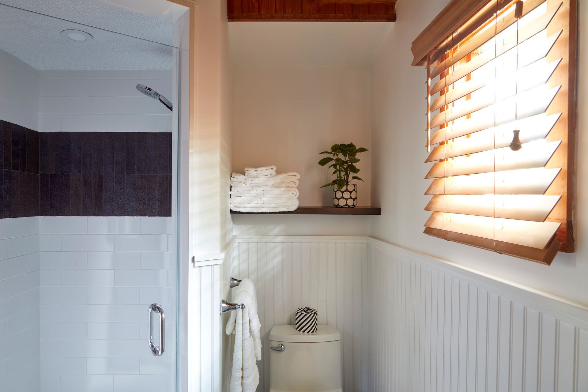 Small bathroom with shower, toilet, towel shelf, and window blinds letting in warm light
