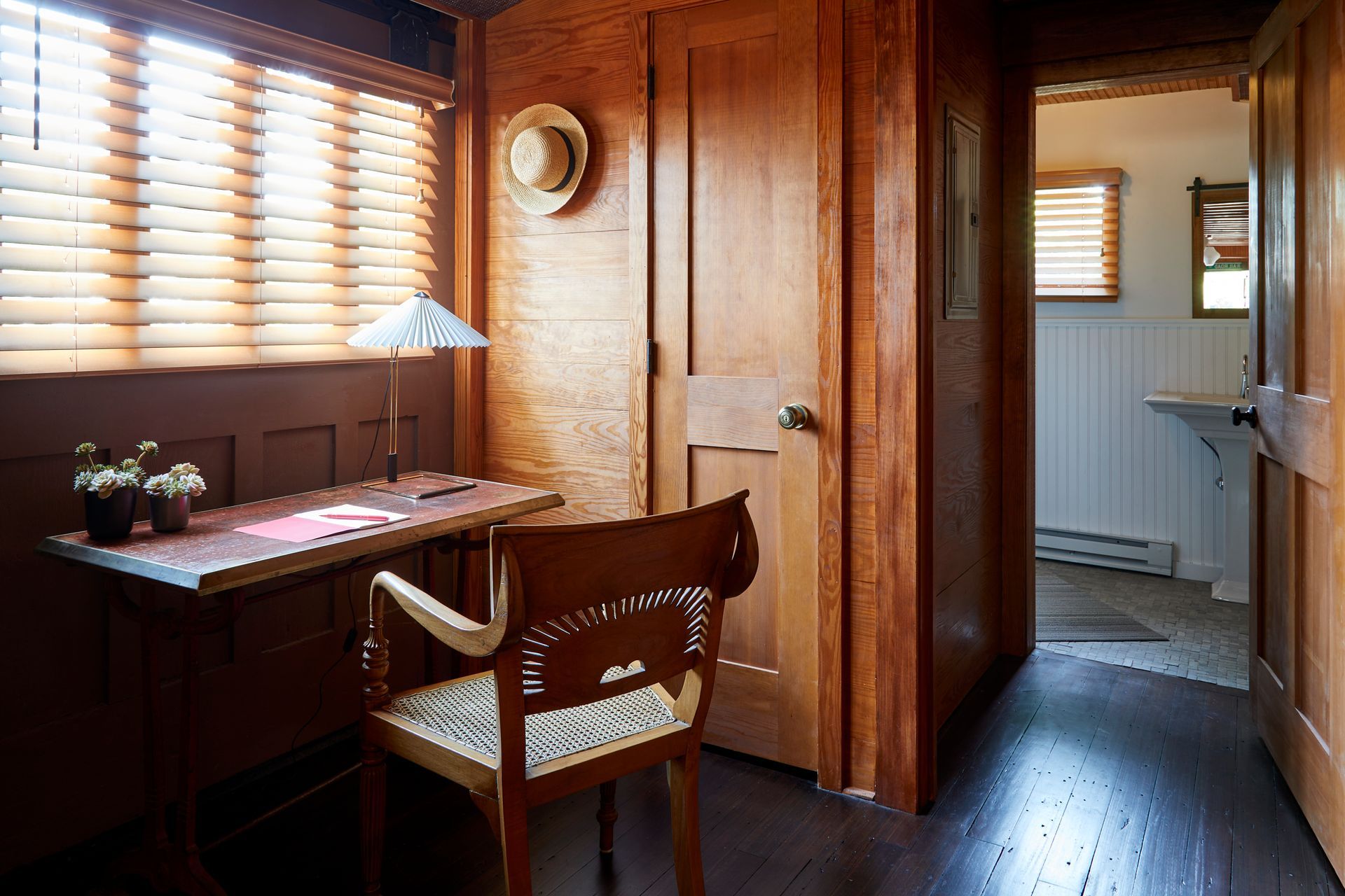 Wood-paneled room with desk, chair, window blinds, and a doorway to a bright adjoining room