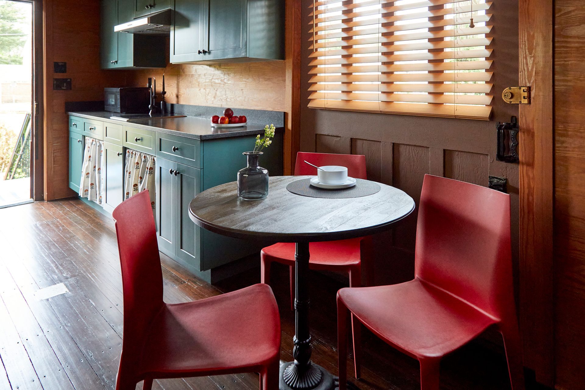 Modern kitchen nook with a round table and two red chairs beside a window and cabinets