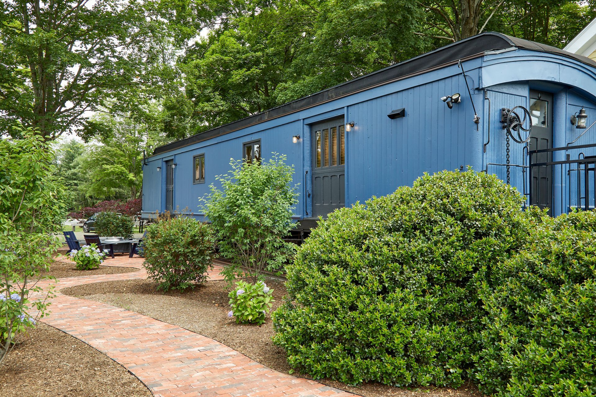 Blue mobile home beside a brick path, surrounded by green shrubs and trees