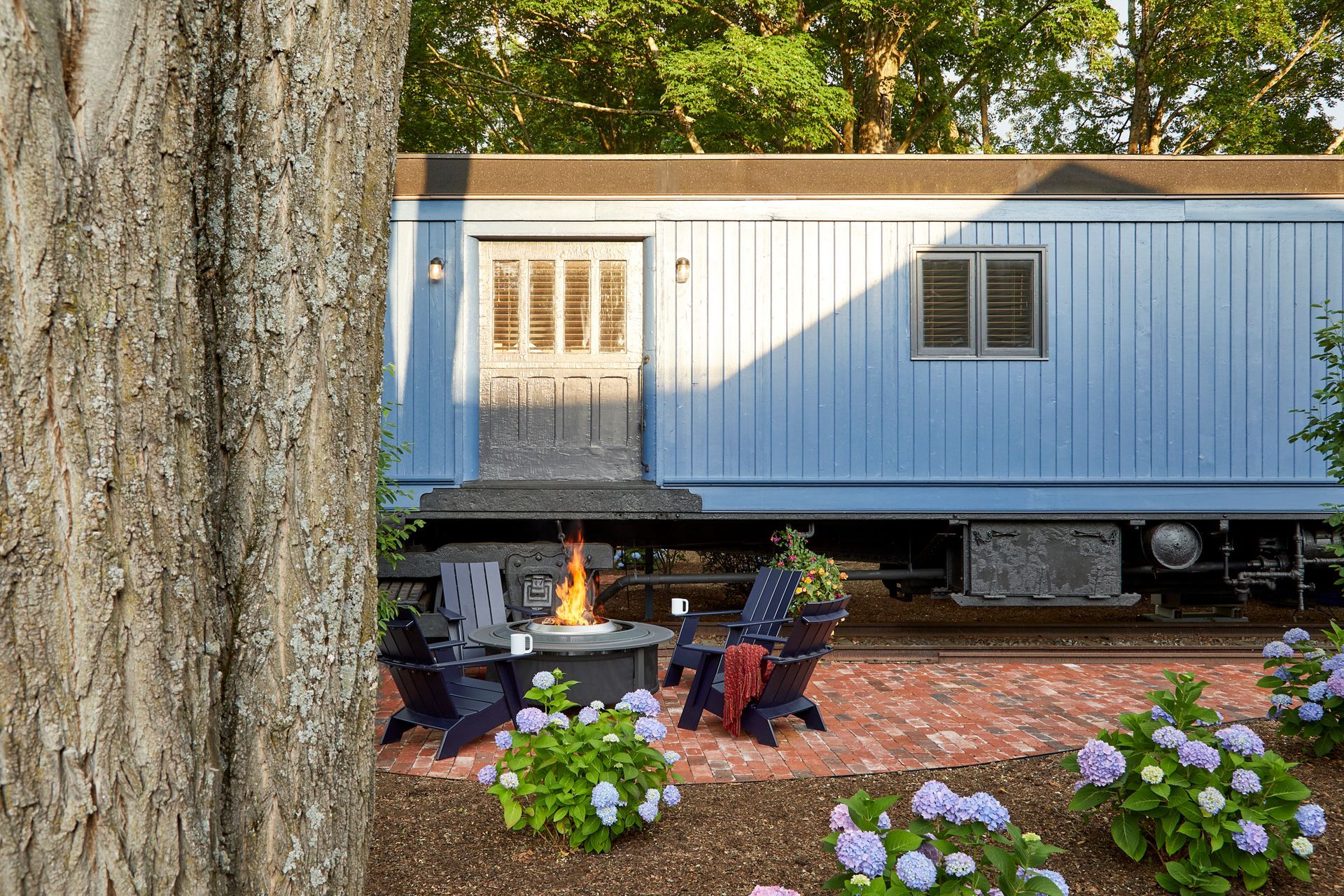 Blue tiny home with porch fire pit and flowers beside a tree in a wooded yard