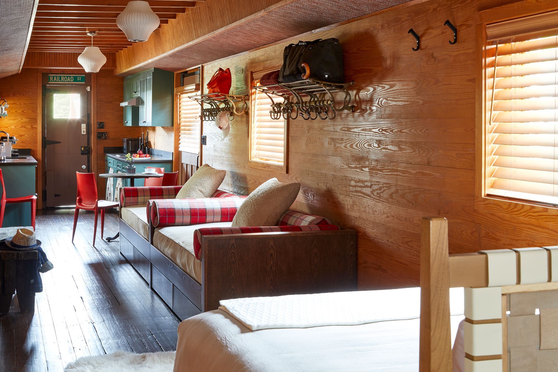 Cozy cabin bedroom with wood-paneled walls, a bed, couch, red chair, and sunlight through blinds