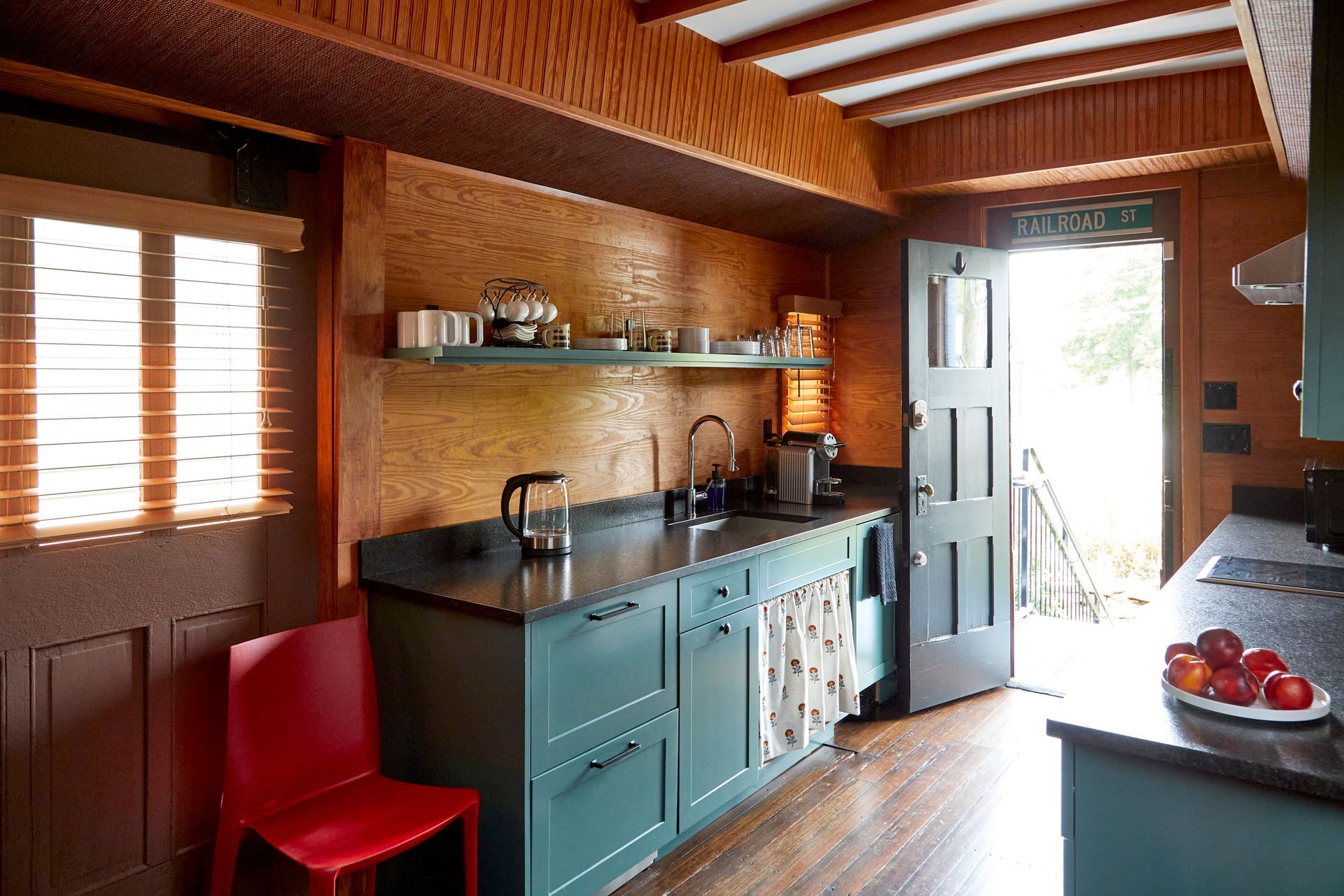 Small rustic kitchen with wood walls, teal cabinets, red chair, and sunlight through windows.