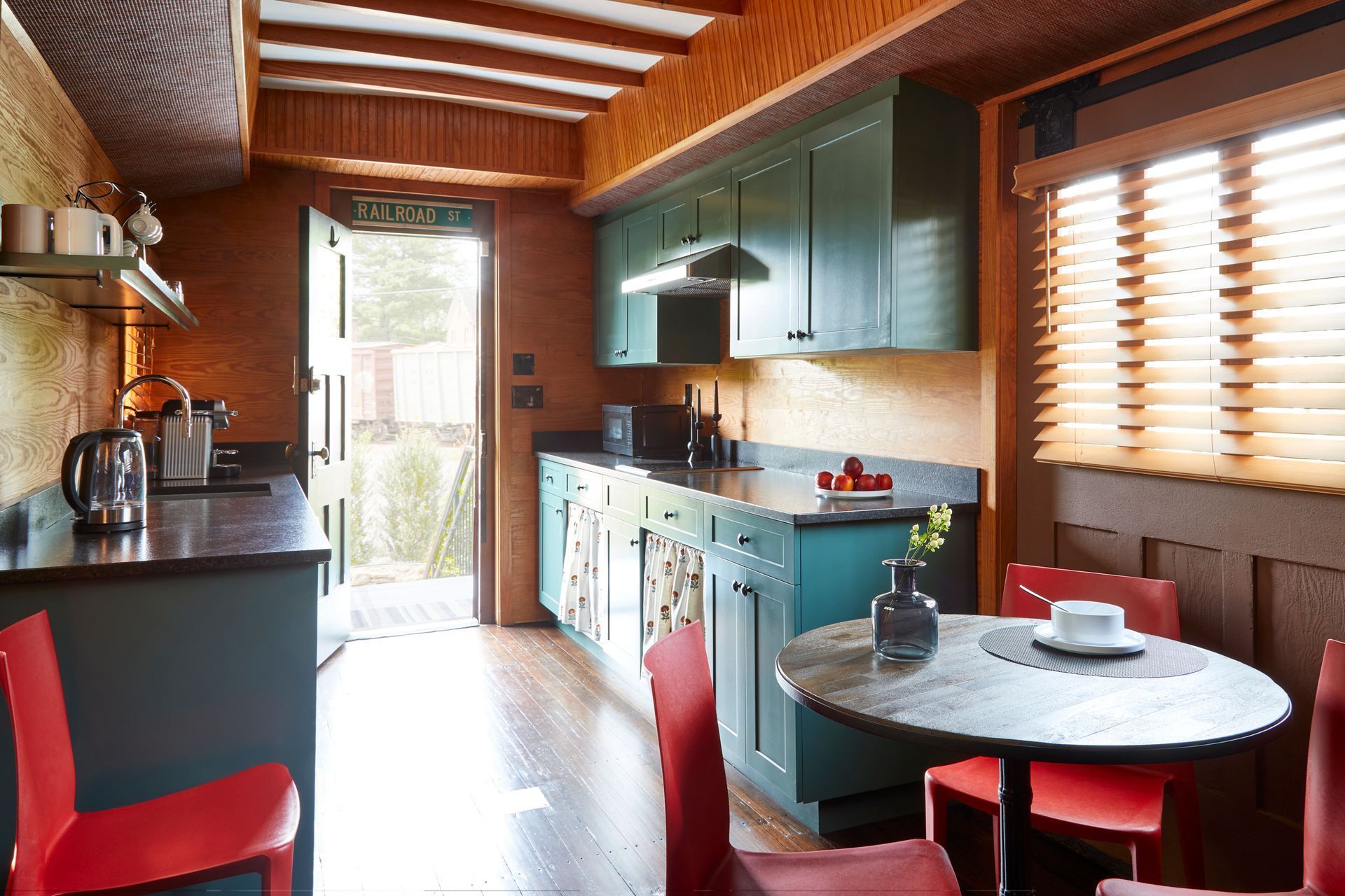 Bright kitchen with teal cabinets, wooden ceiling, round table, and sunlight through blinds.