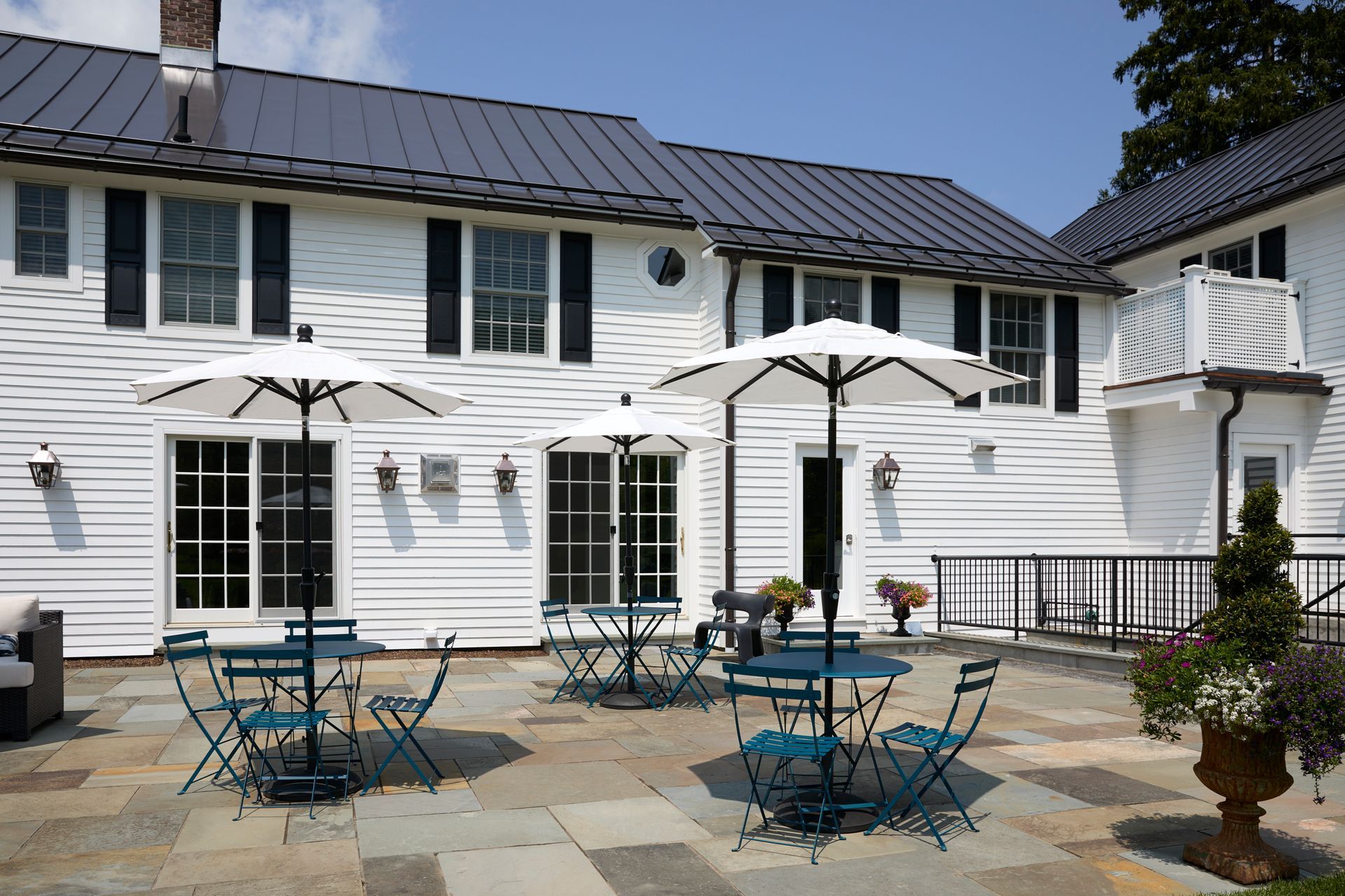 White patio with blue tables and umbrellas in front of a white house with black shutters and French doors