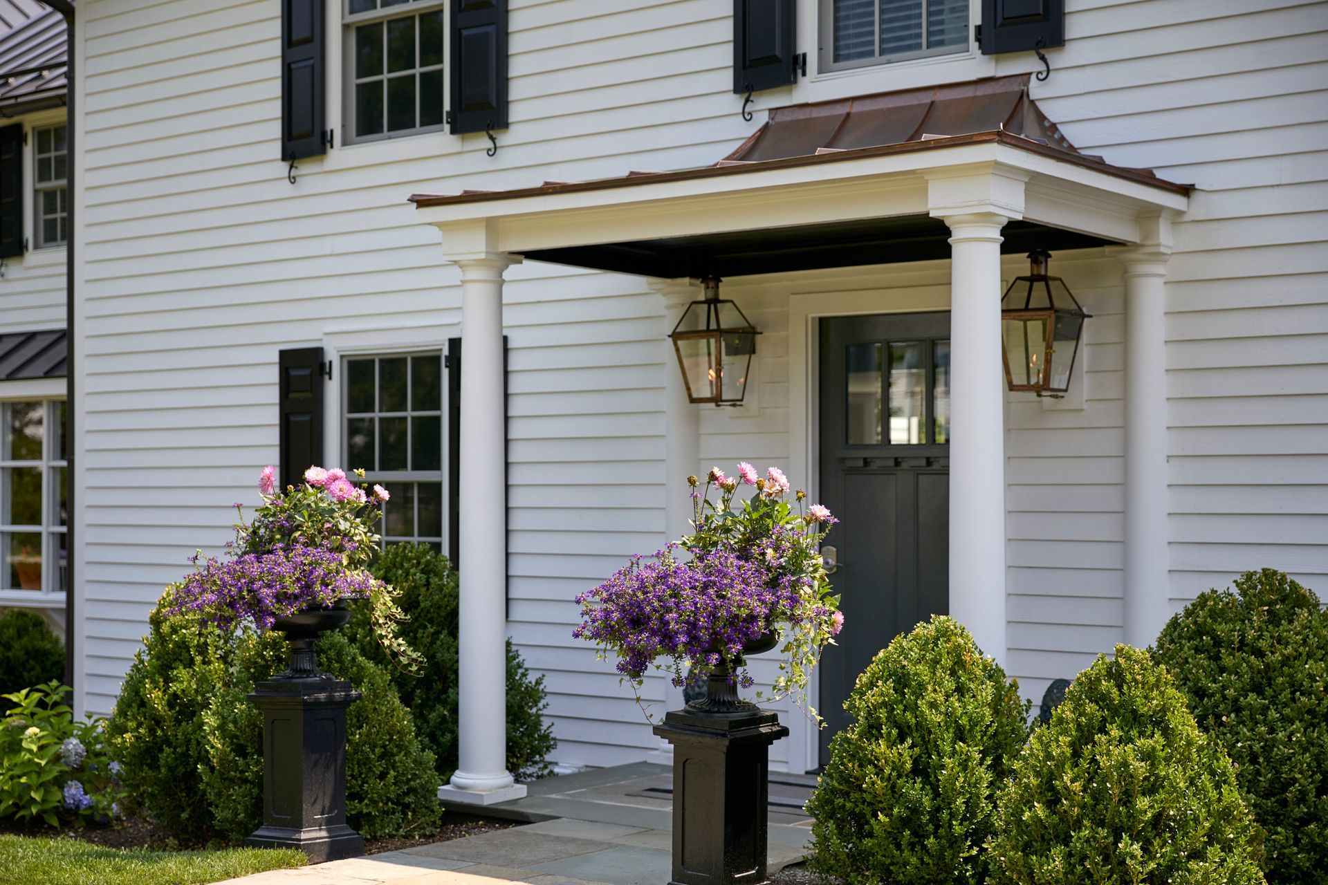 White house entry with porch columns, hanging lanterns, and purple potted flowers by the front door