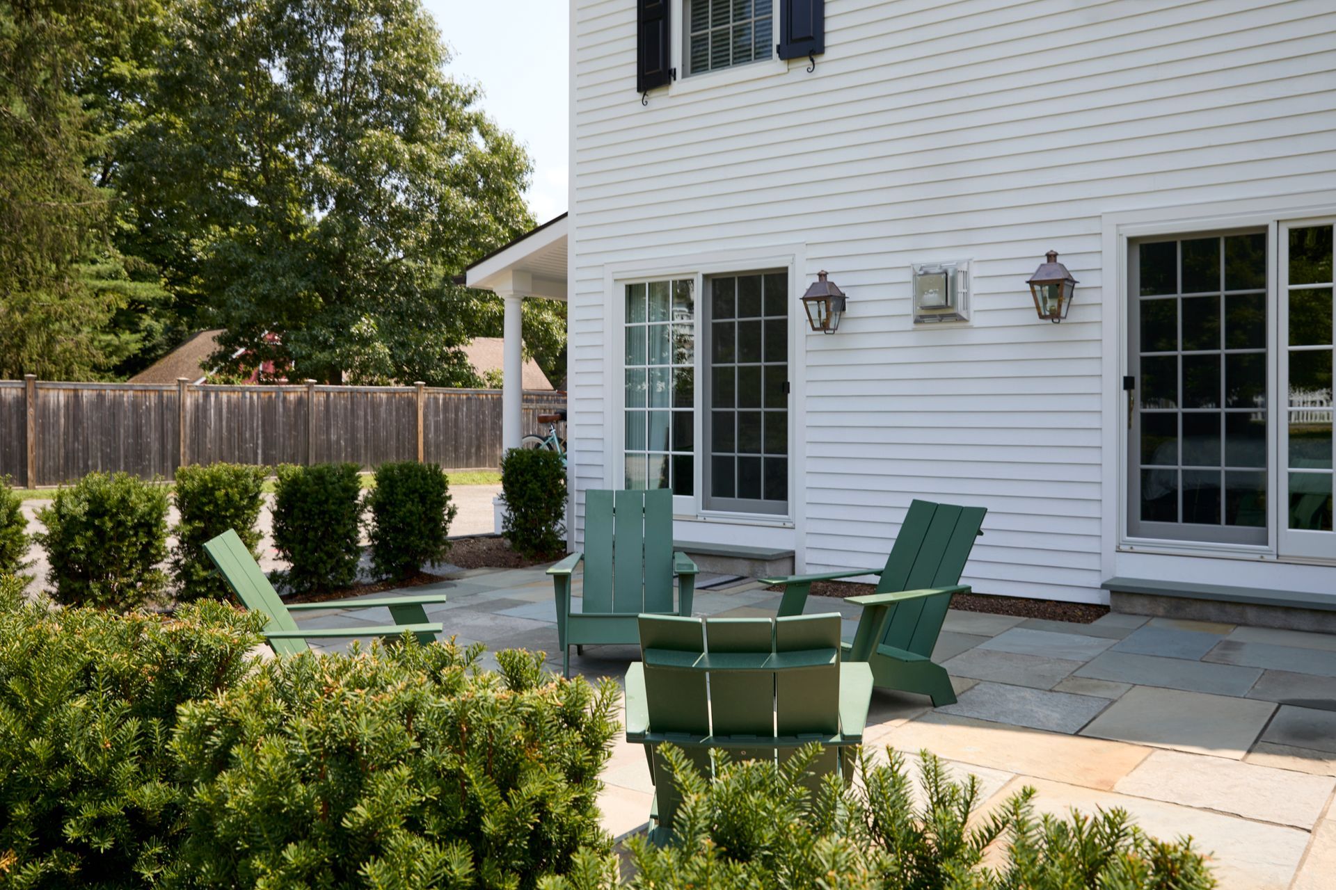 Back patio with green chairs and table beside a white house and wooden fence.