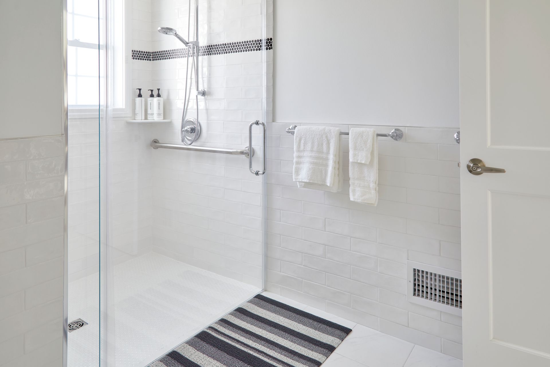 Bright white tiled bathroom shower with glass door, towel bars, and striped bath mat