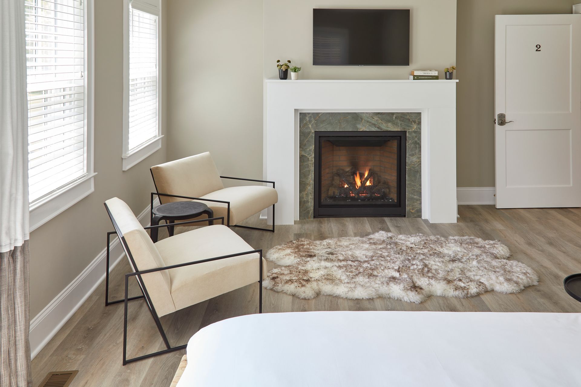 Modern bedroom with fireplace, TV, beige chairs, shag rug, and gray wood flooring