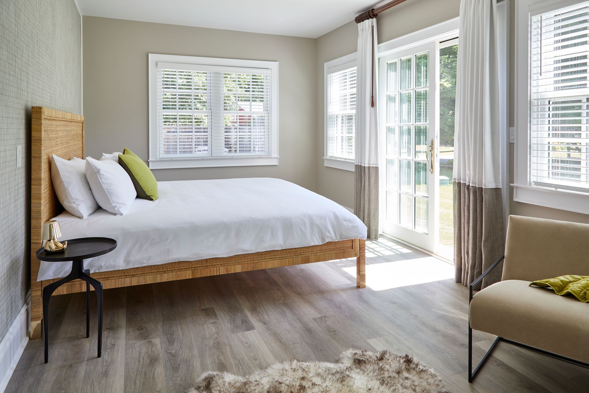 Bright bedroom with a white bed, windows, sheer curtains, and a beige chair on a light wood floor.