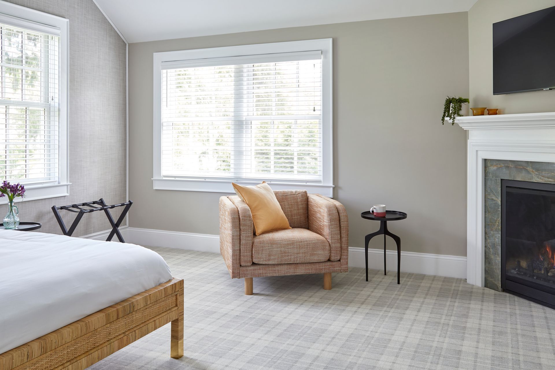 Bright bedroom with a bed, wicker chair, large window, and fireplace on light carpet.